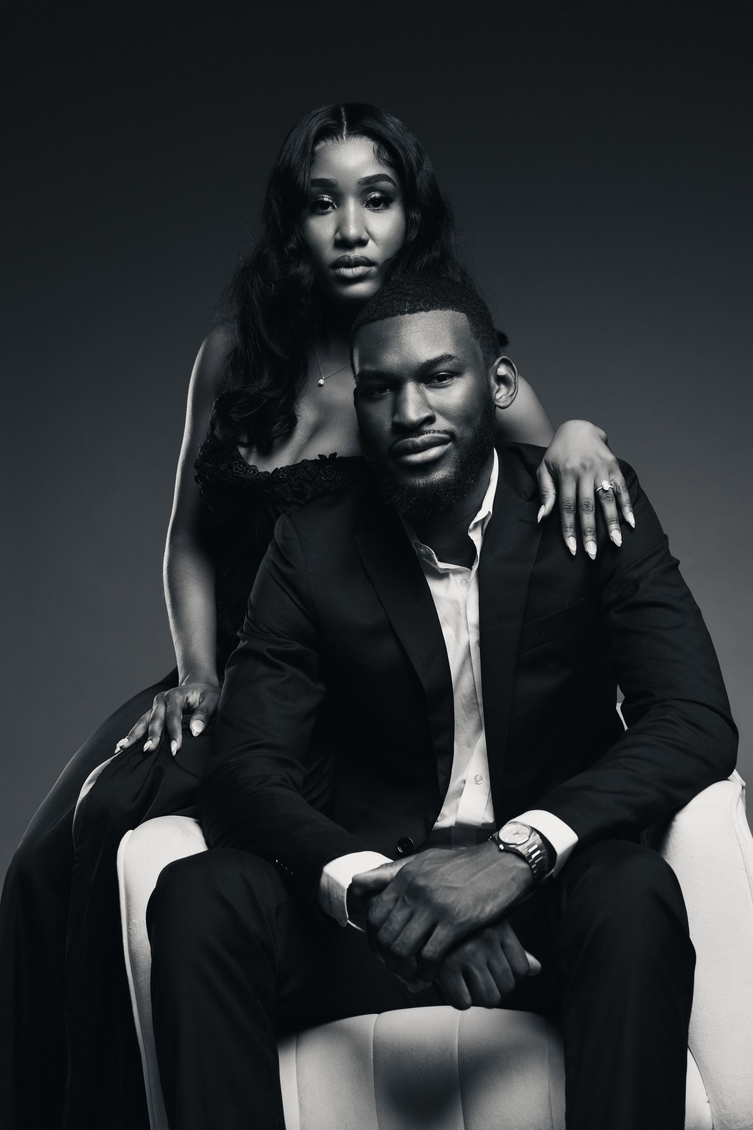 Black and white portrait of stylish couple, man in suit seated in white chair, woman in black dress standing behind him.