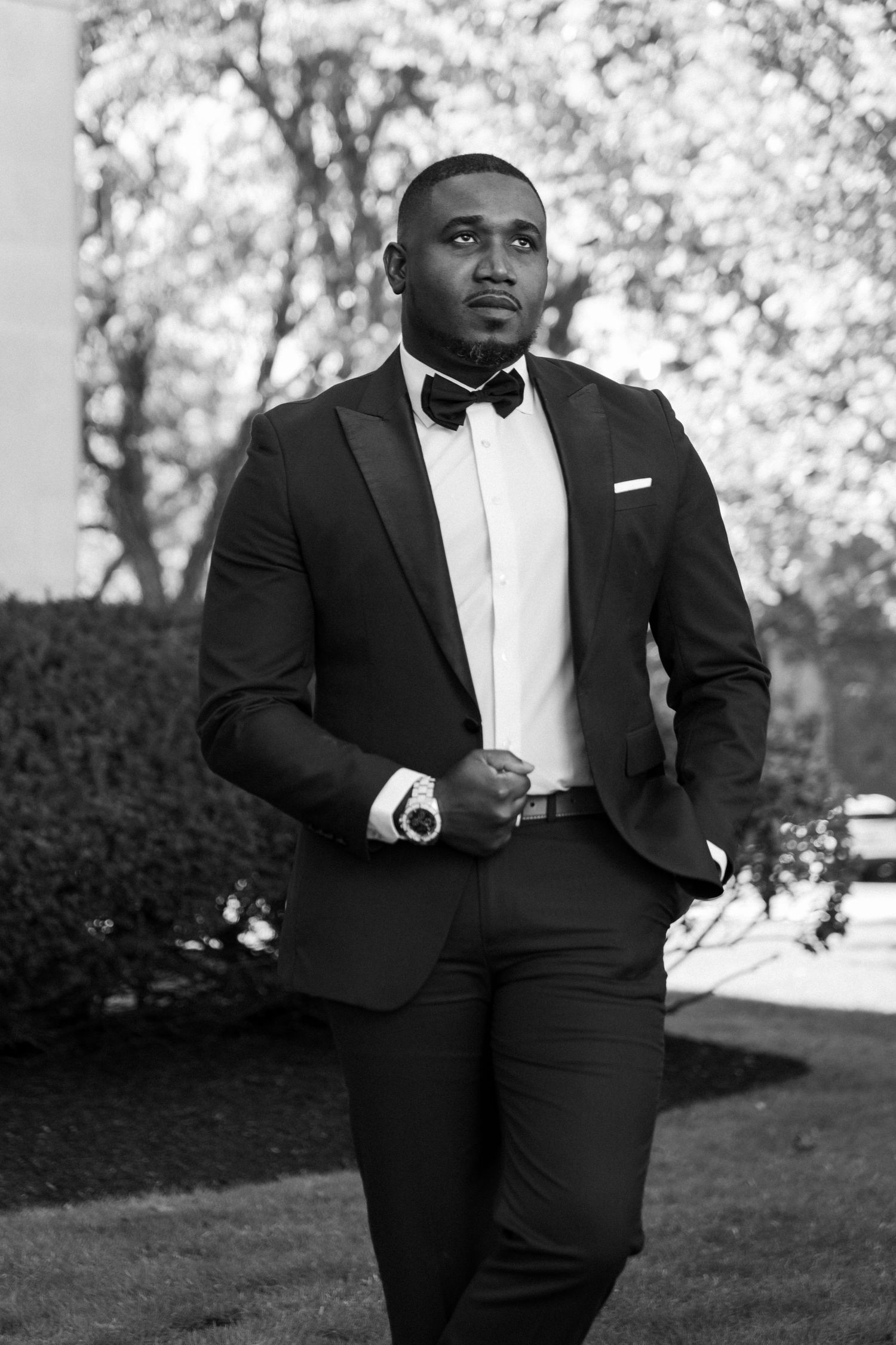 Elegant groom in black tuxedo with bow tie poses confidently outdoors in black and white portrait photo.