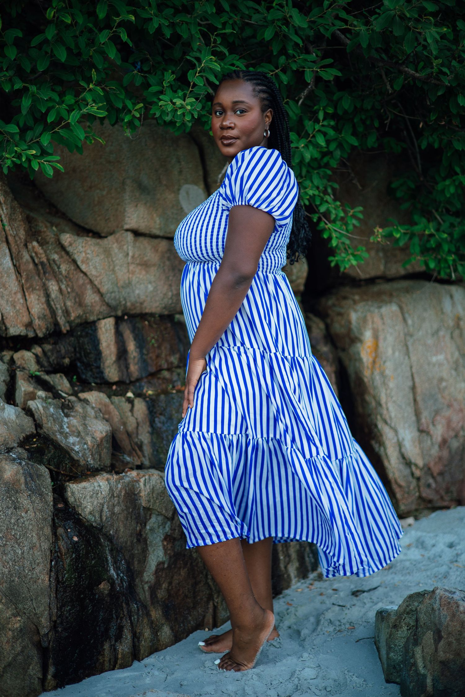 Woman in blue and white striped midi dress poses gracefully on rocky beach with lush green foliage backdrop.