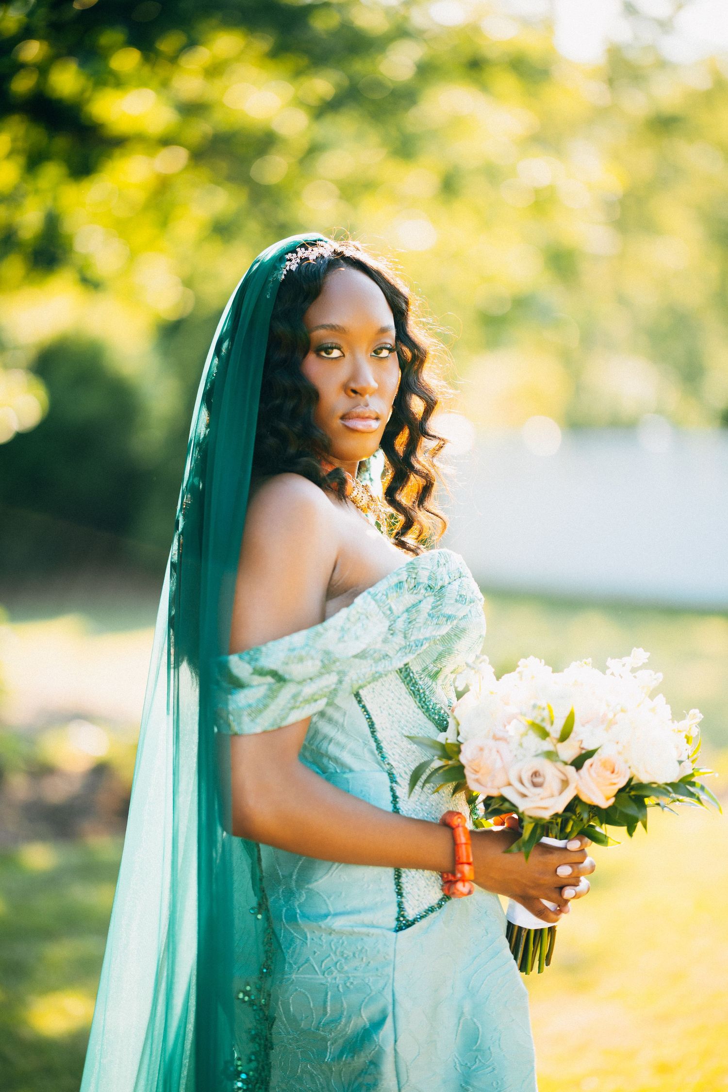 A Black bride in a teal off-shoulder gown and veil holds a white bouquet outdoors in soft sunlight.