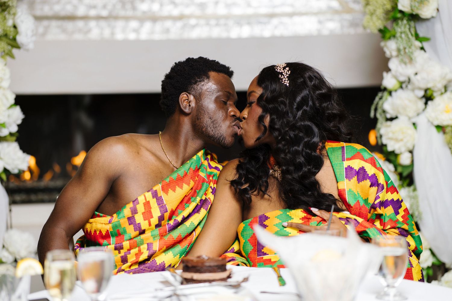 Couple in traditional Kente cloth attire sharing a kiss at a decorated wedding reception table.