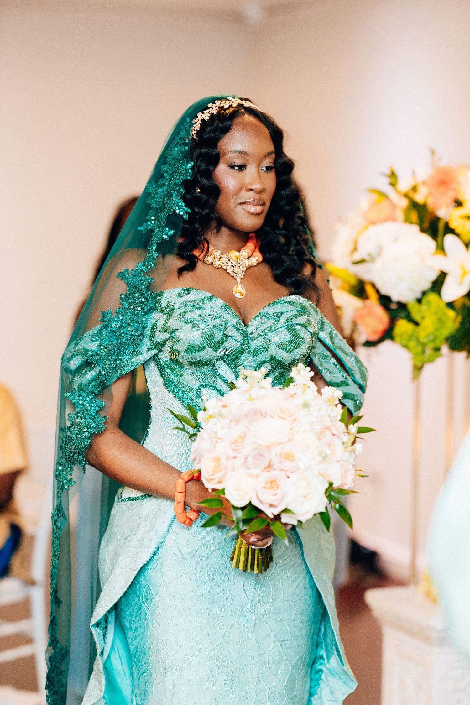 A bride in a teal lace gown and matching veil holds a white floral bouquet, adorned with gold jewelry.