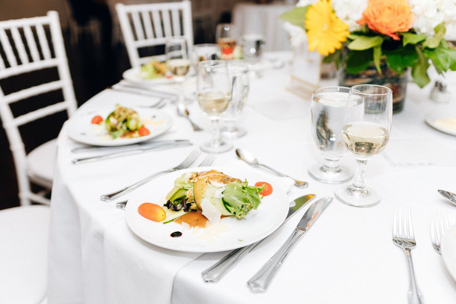 Formal dining table set with white plates of fresh salad, silverware, wine glasses, and colorful flower arrangement.