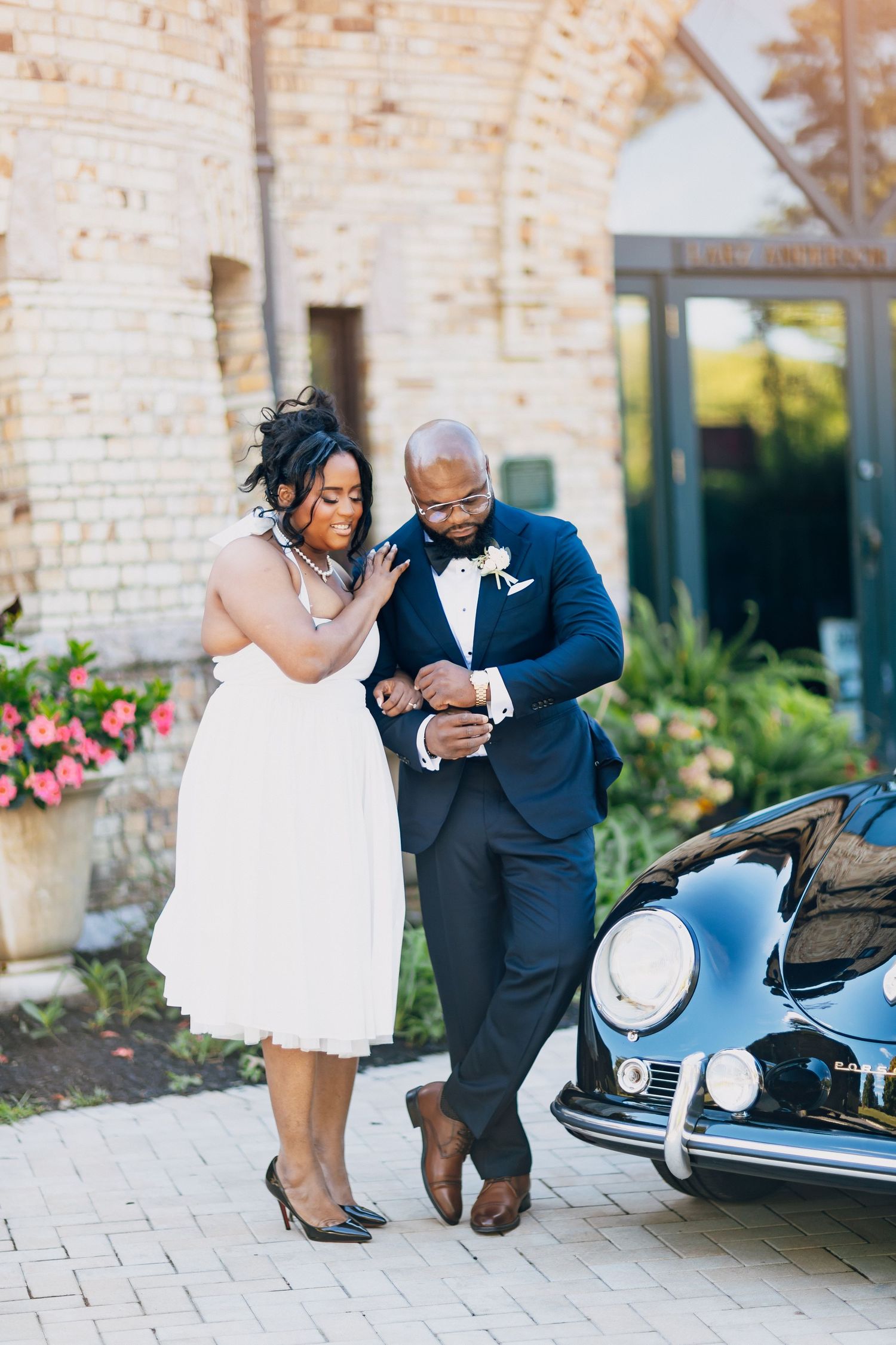 A bride and groom share a joyful moment beside a vintage black car outside a stone building.