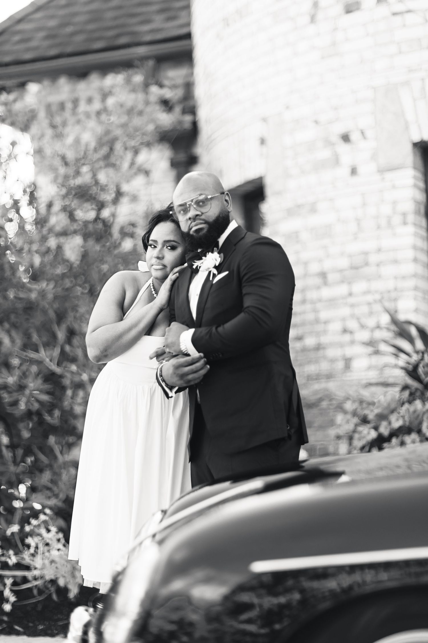 Black and white portrait of a bride and groom embracing near a vintage car outside a brick building.