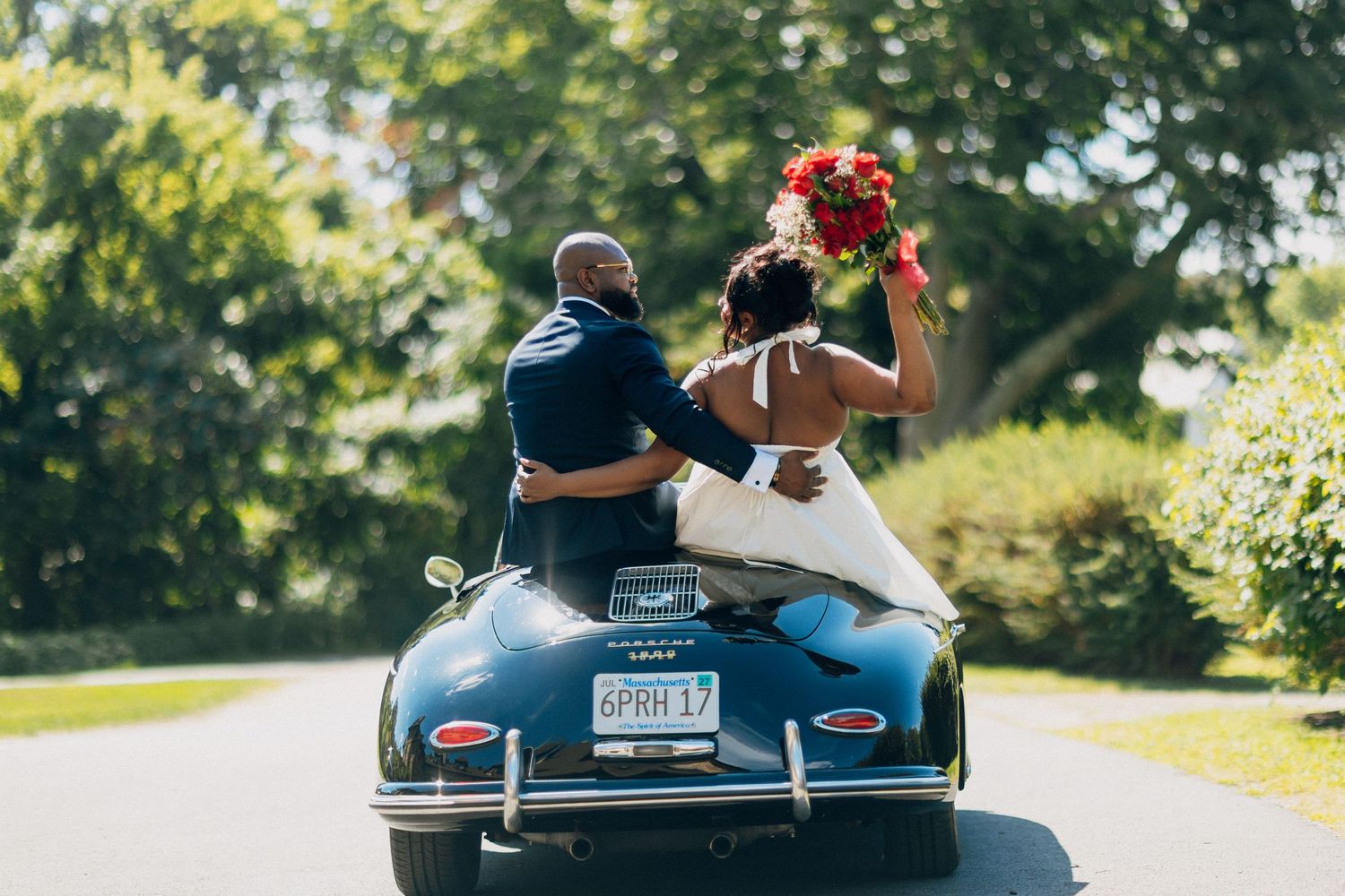 Black bride raising colorful bouquet while riding with groom in a classic vintage Porsche convertible.