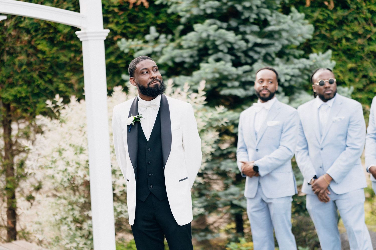 Groom in white tuxedo jacket stands at outdoor wedding altar with two groomsmen in light blue suits behind him.
