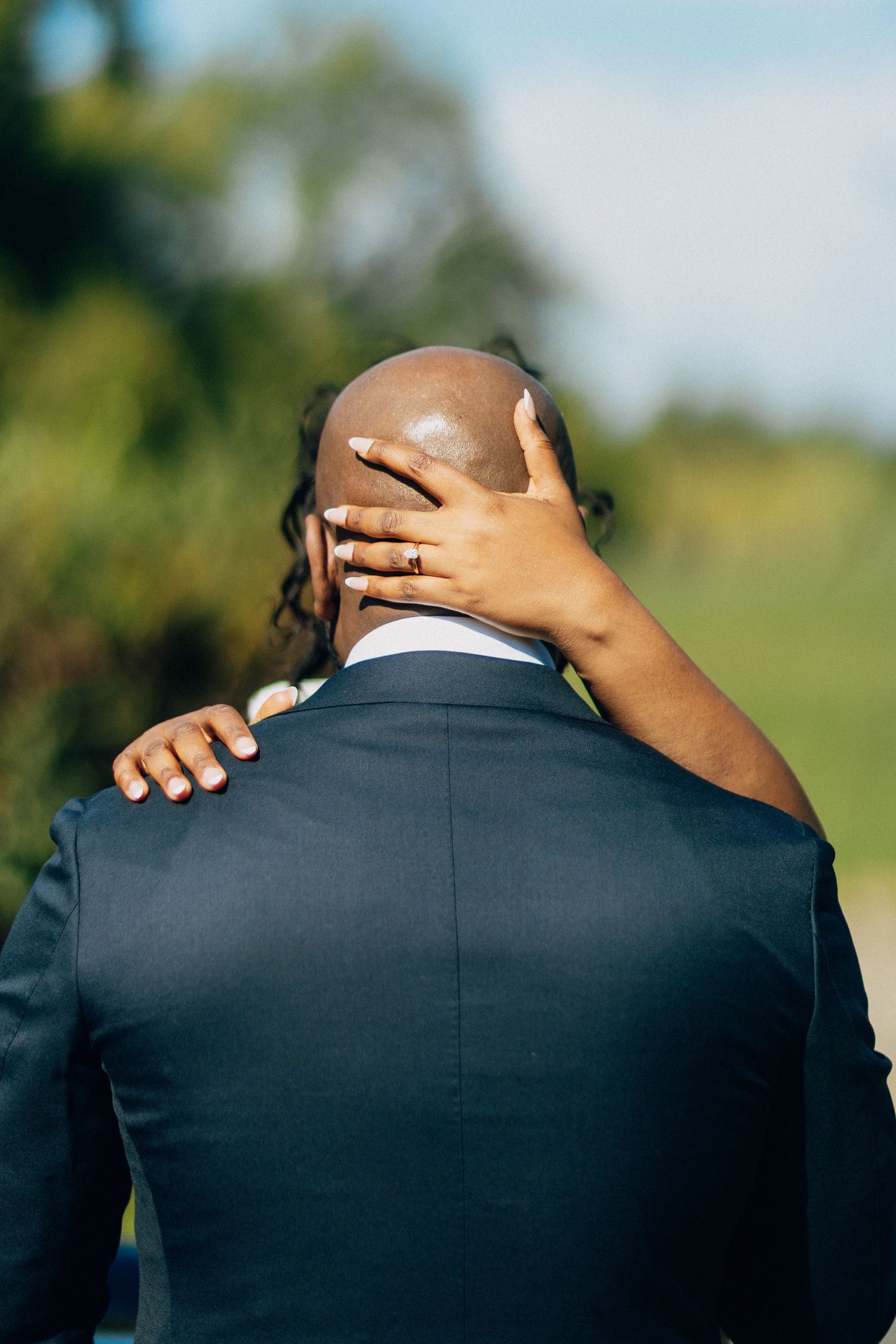 A bride places her hand on the back of her grooms head during an outdoor wedding celebration.