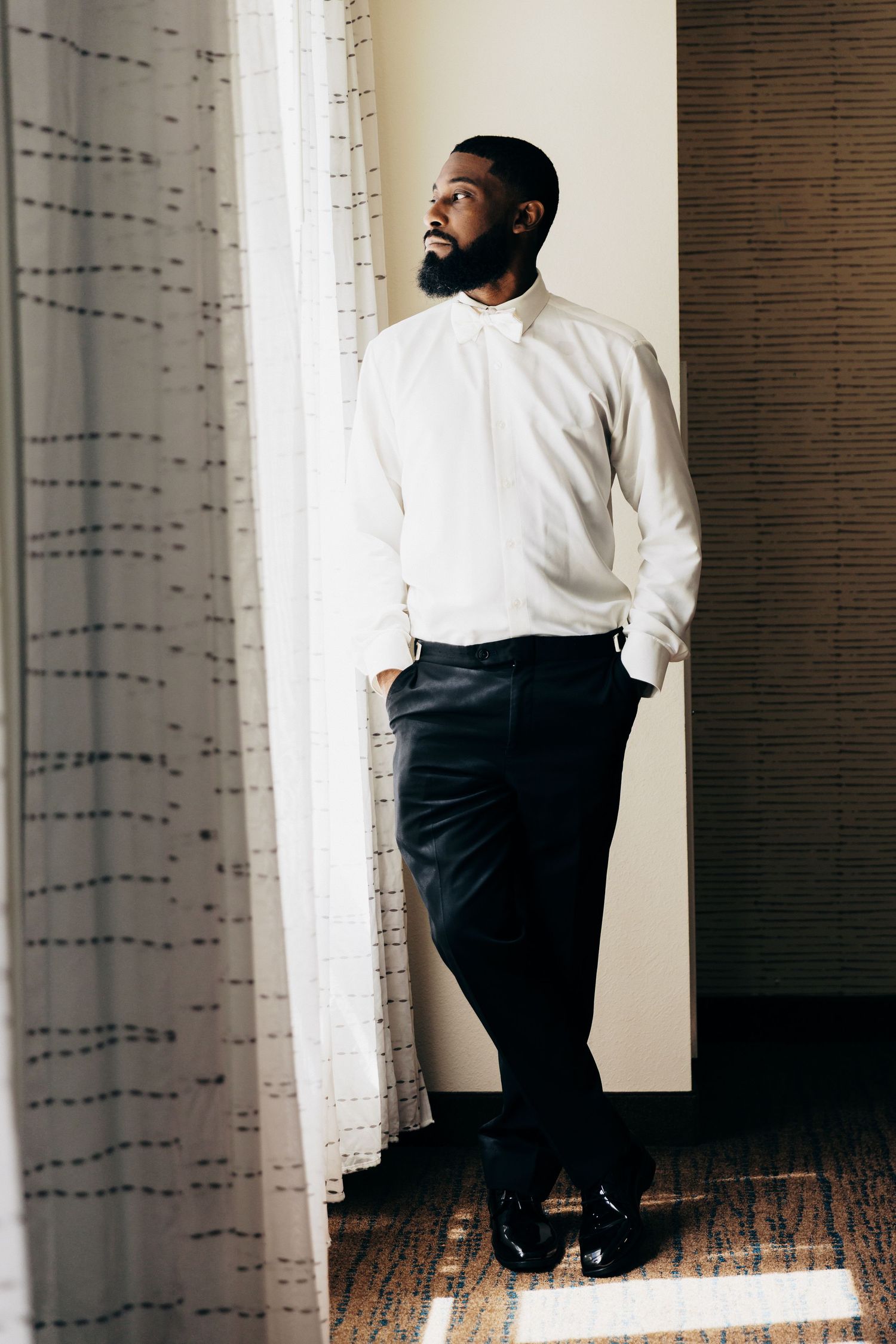 A well-dressed Black man in a white dress shirt and black trousers stands by a window, gazing thoughtfully into the distance.