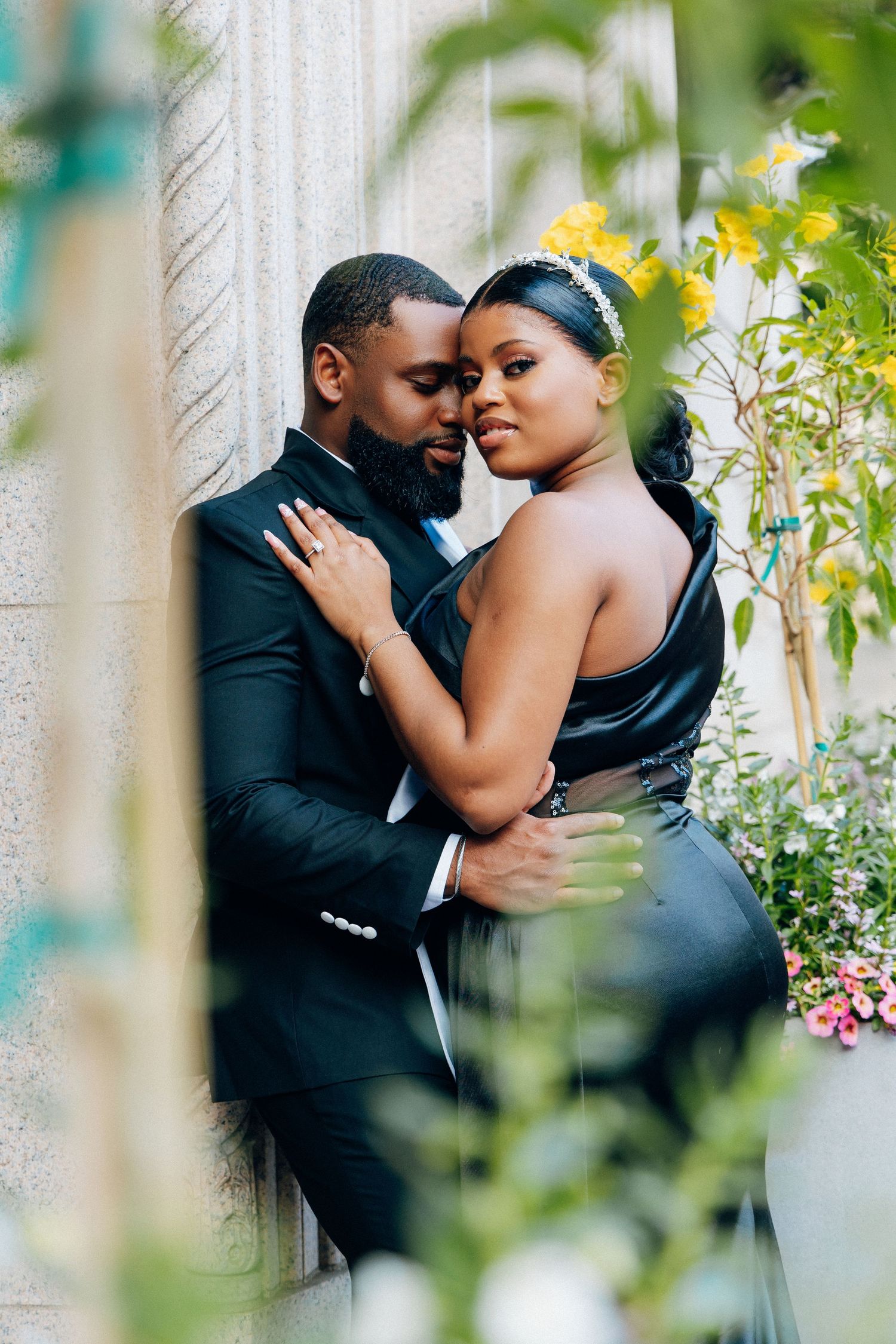 A couple in elegant black formal wear shares a romantic embrace against a stone wall surrounded by lush greenery.