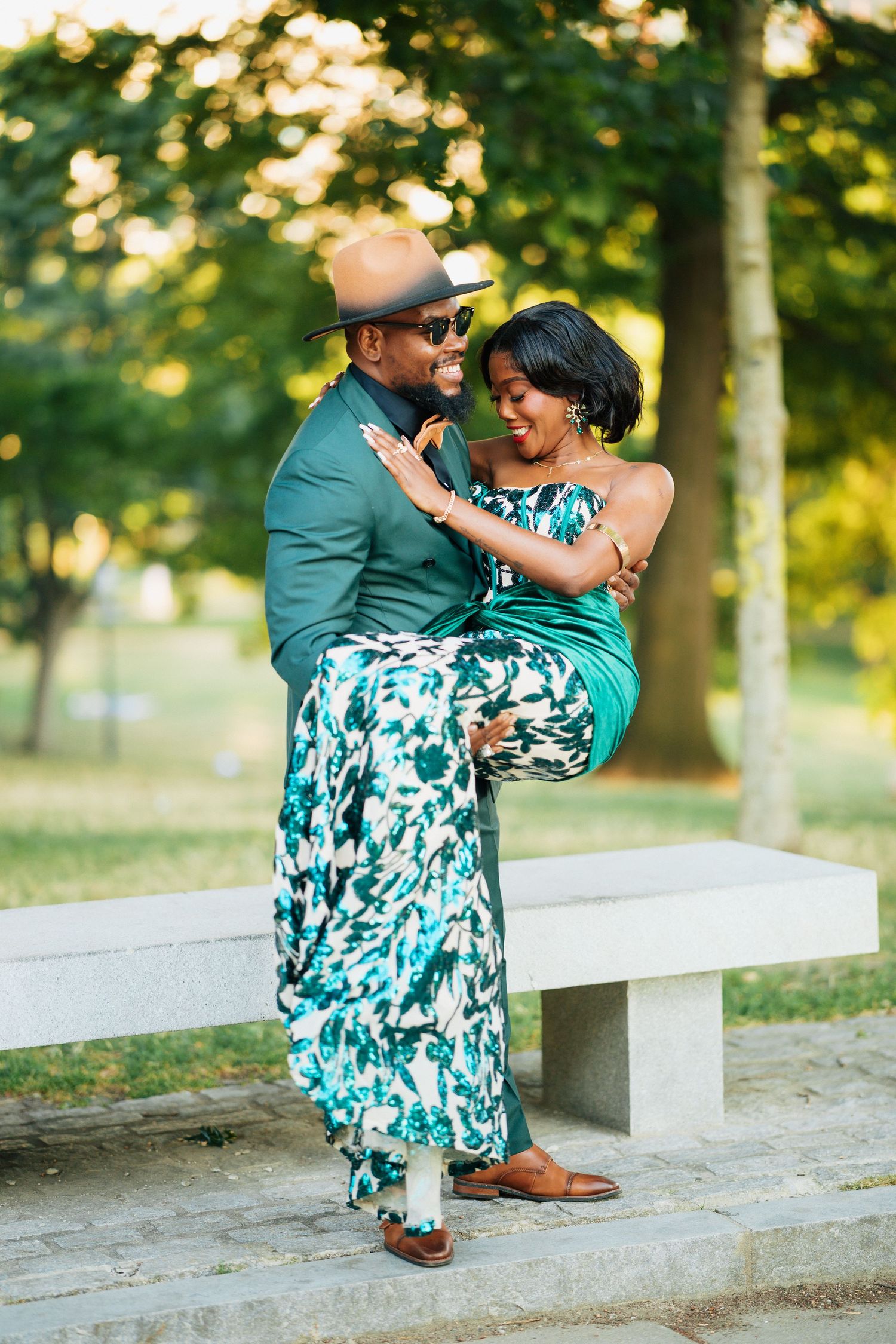 Stylish couple in green outfits sharing a romantic moment on a park bench, man lifting woman playfully.