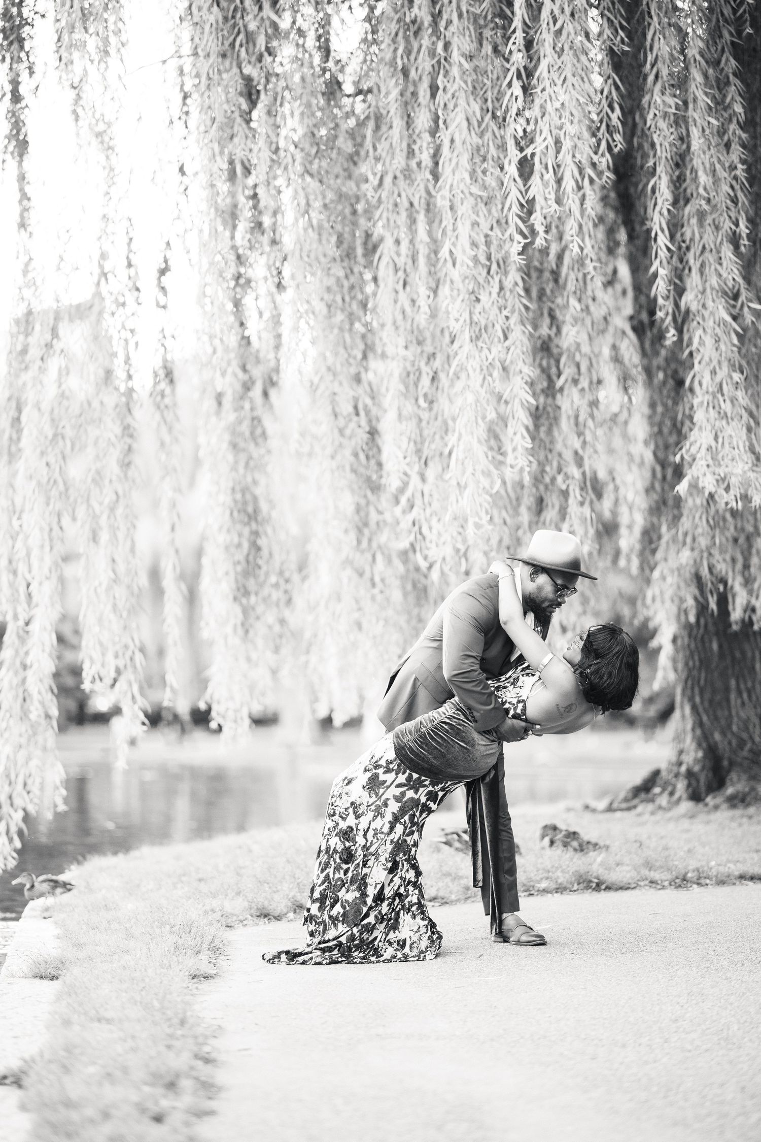 Black and white photo of couple sharing a romantic dip kiss under a weeping willow tree by a lake.