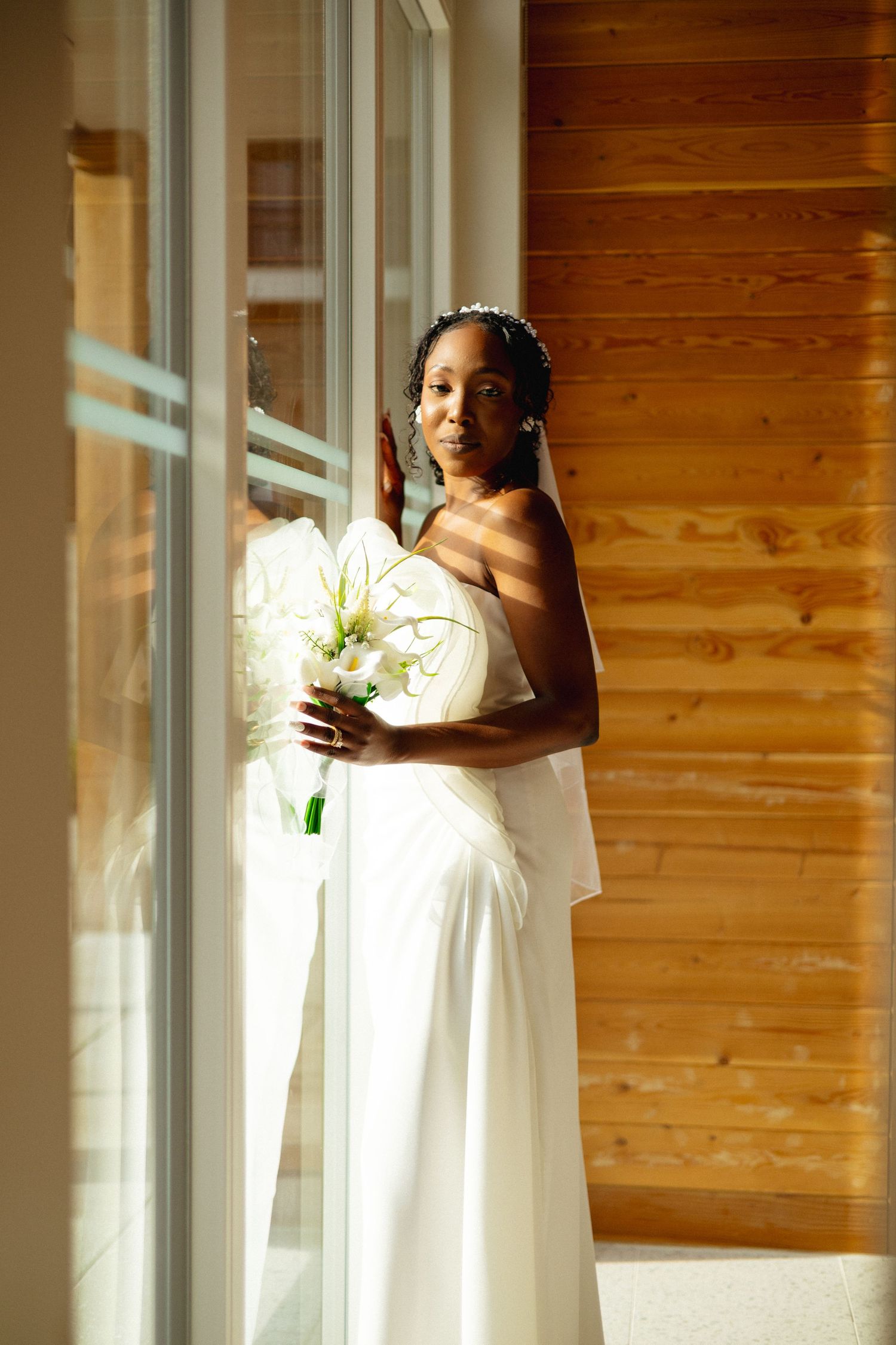 Beautiful Black bride in white strapless wedding dress holds calla lilies beside a sunlit window with wooden wall.