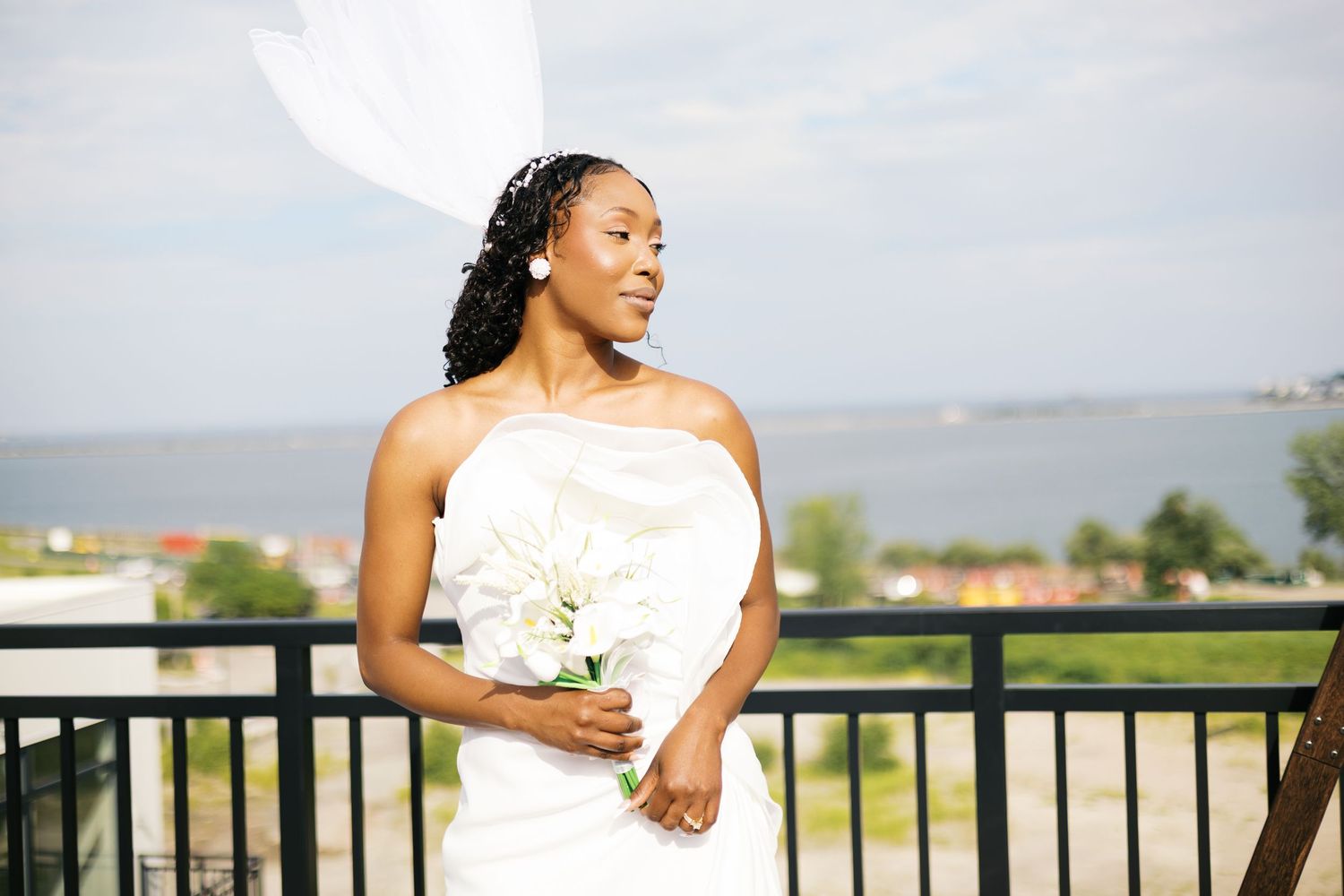 Elegant bride in strapless white gown holding bouquet on balcony overlooking water on her wedding day.