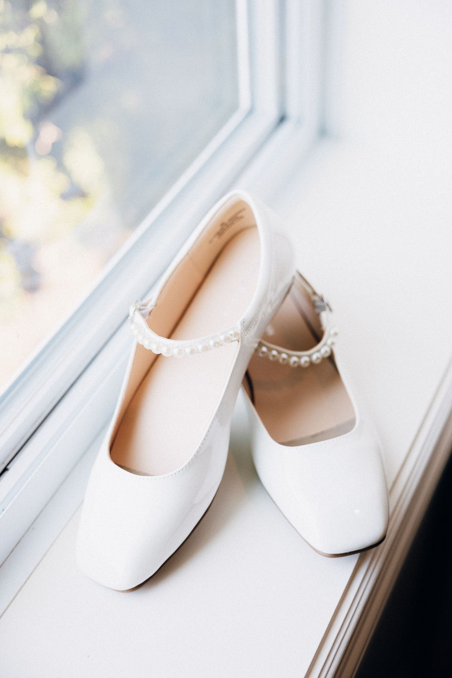 White bridal heels with pearl ankle strap detail displayed on a window sill in natural light.