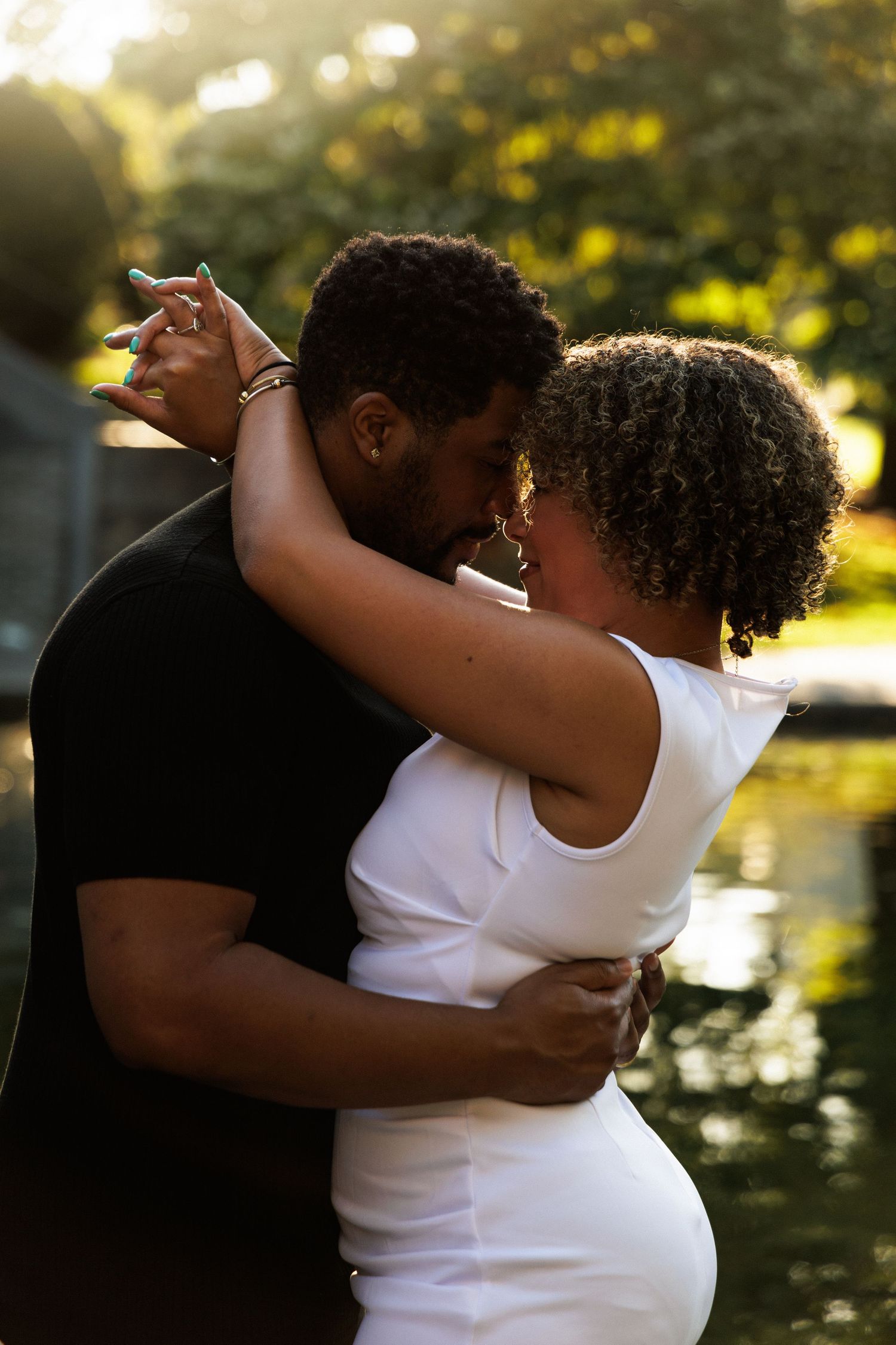 A couple embraces and kisses near a pond in golden hour sunlight, man in black shirt, woman in white dress.