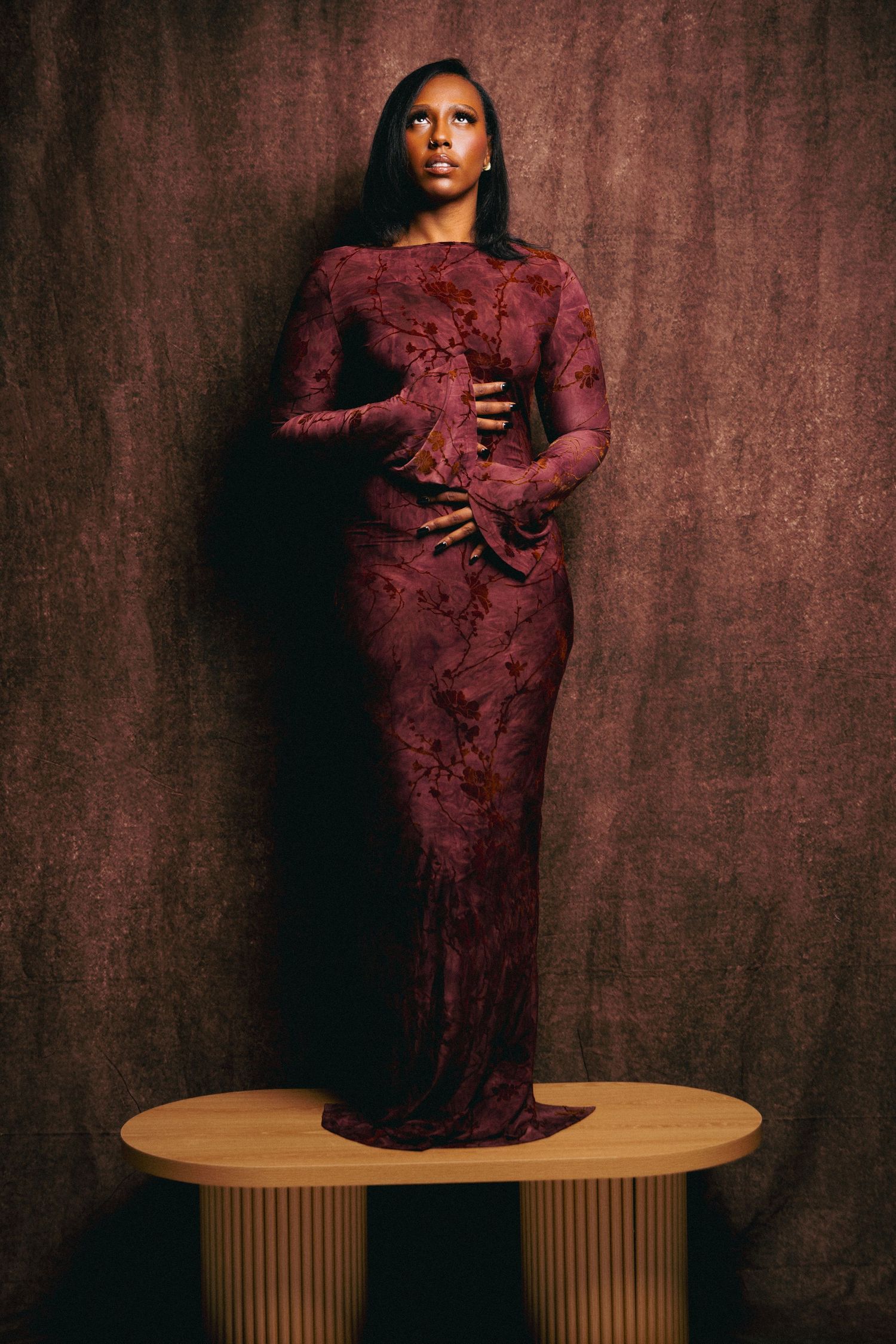 A woman in a burgundy lace gown stands on a wooden platform against a dark brown backdrop.