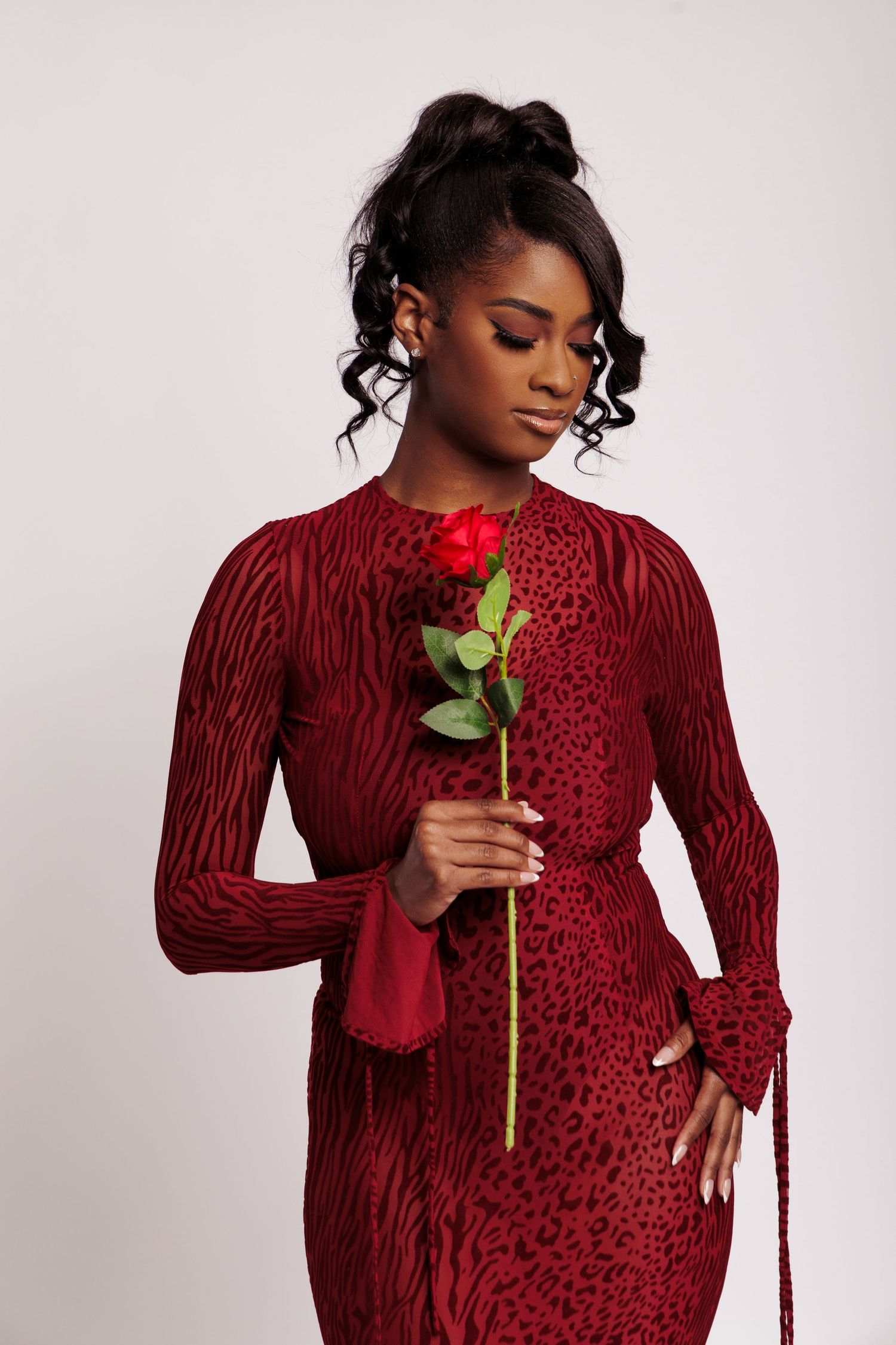 A young Black woman in a red animal print dress holds a single red rose, looking down elegantly against a white background.