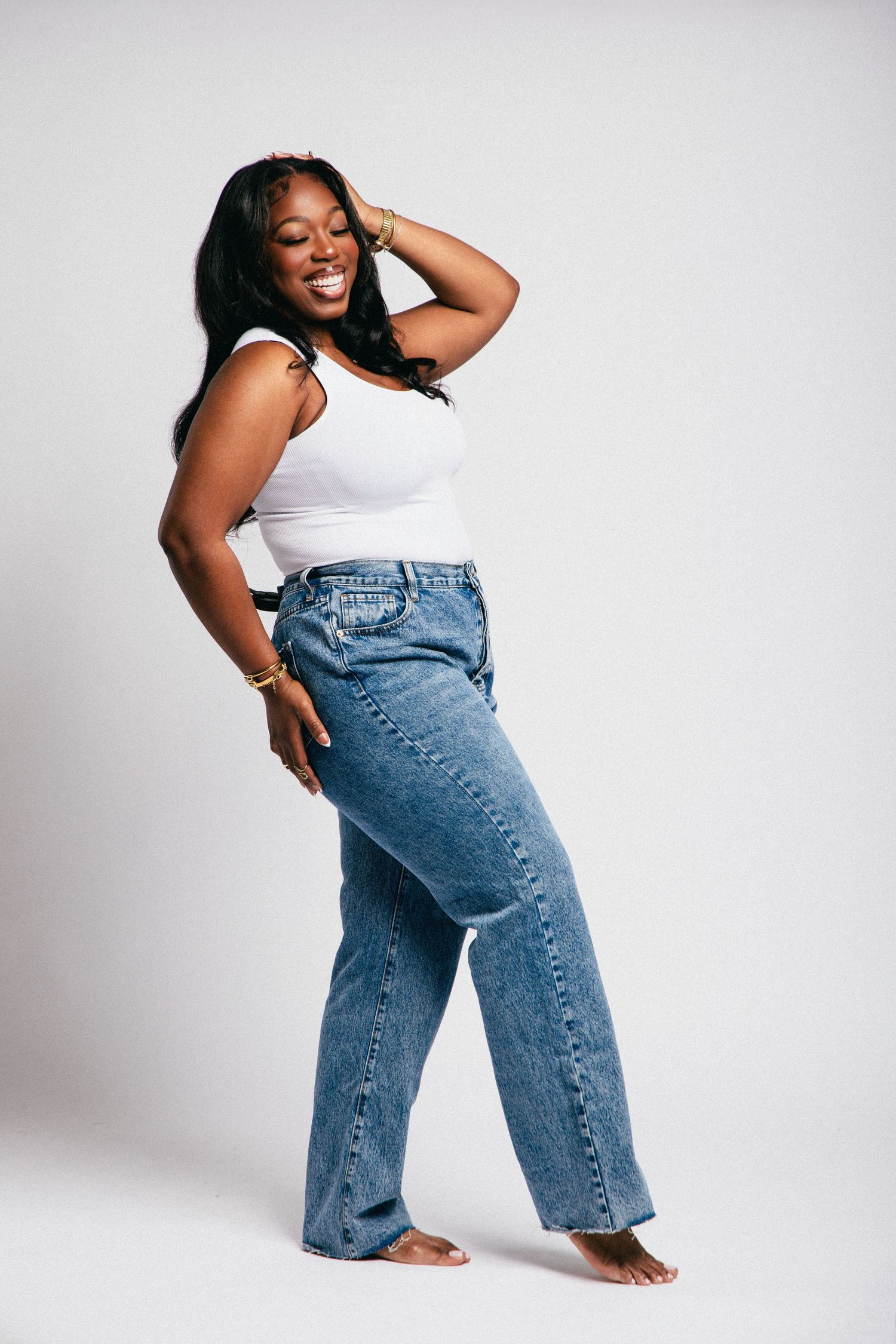 Smiling Black woman posing in white tank top and wide-leg blue jeans, barefoot against a light gray background.
