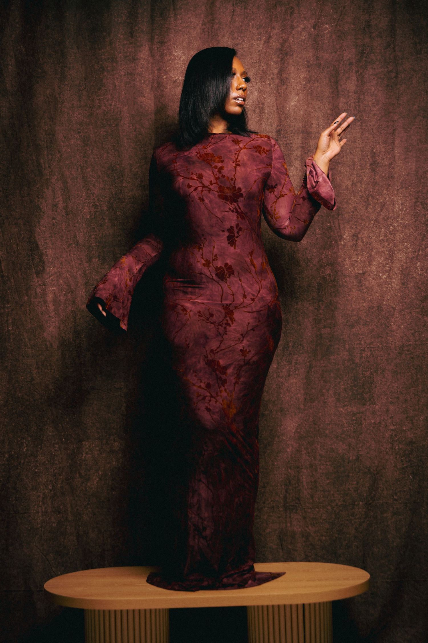 Woman in burgundy floral lace maxi dress posing on pedestal against brown backdrop.