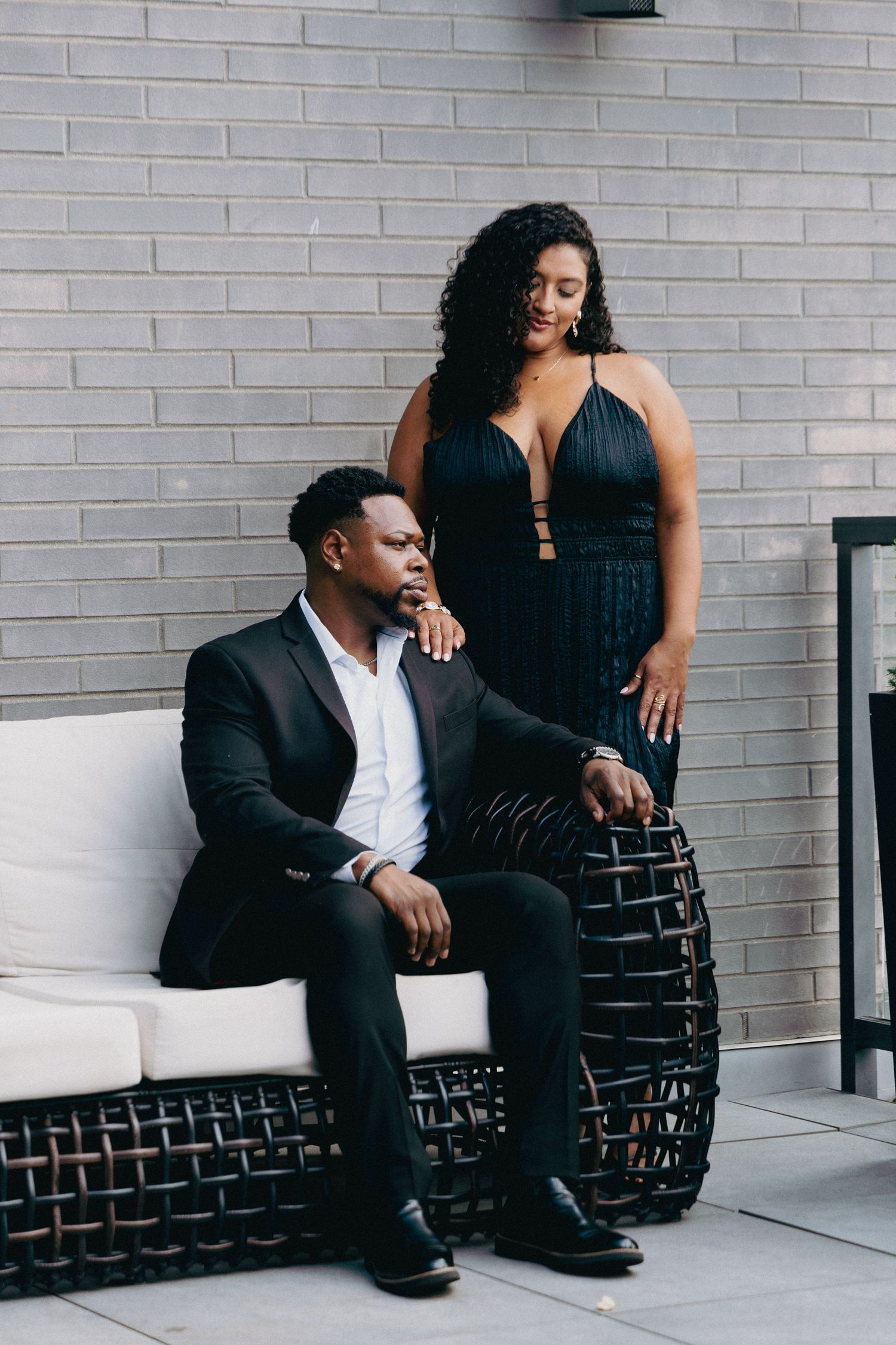 Elegant couple posing outdoors, man in black suit seated on wicker sofa, woman in black halter dress standing beside him.