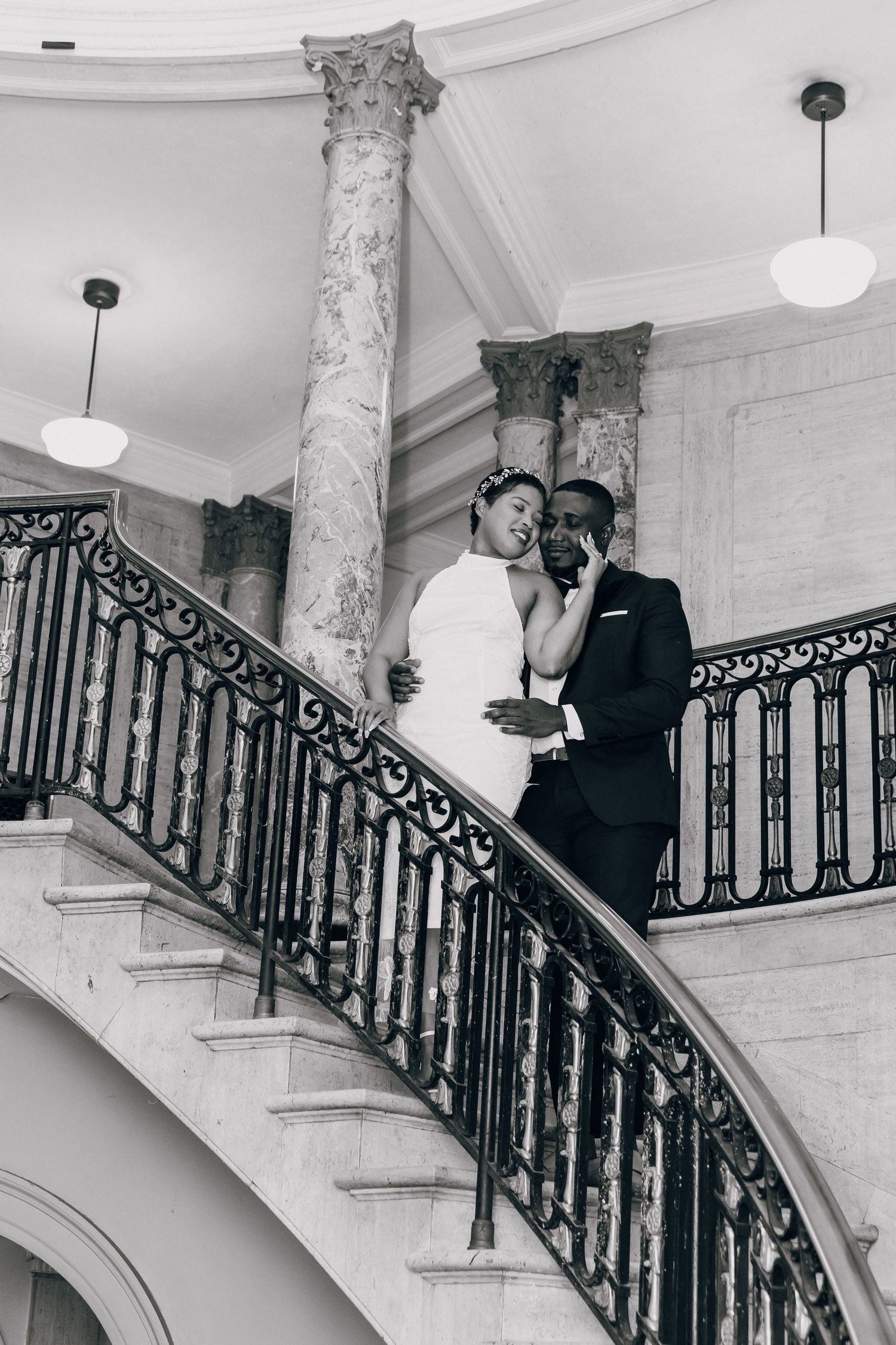 Black and white photo of wedding couple embracing on elegant curved staircase with iron railings.