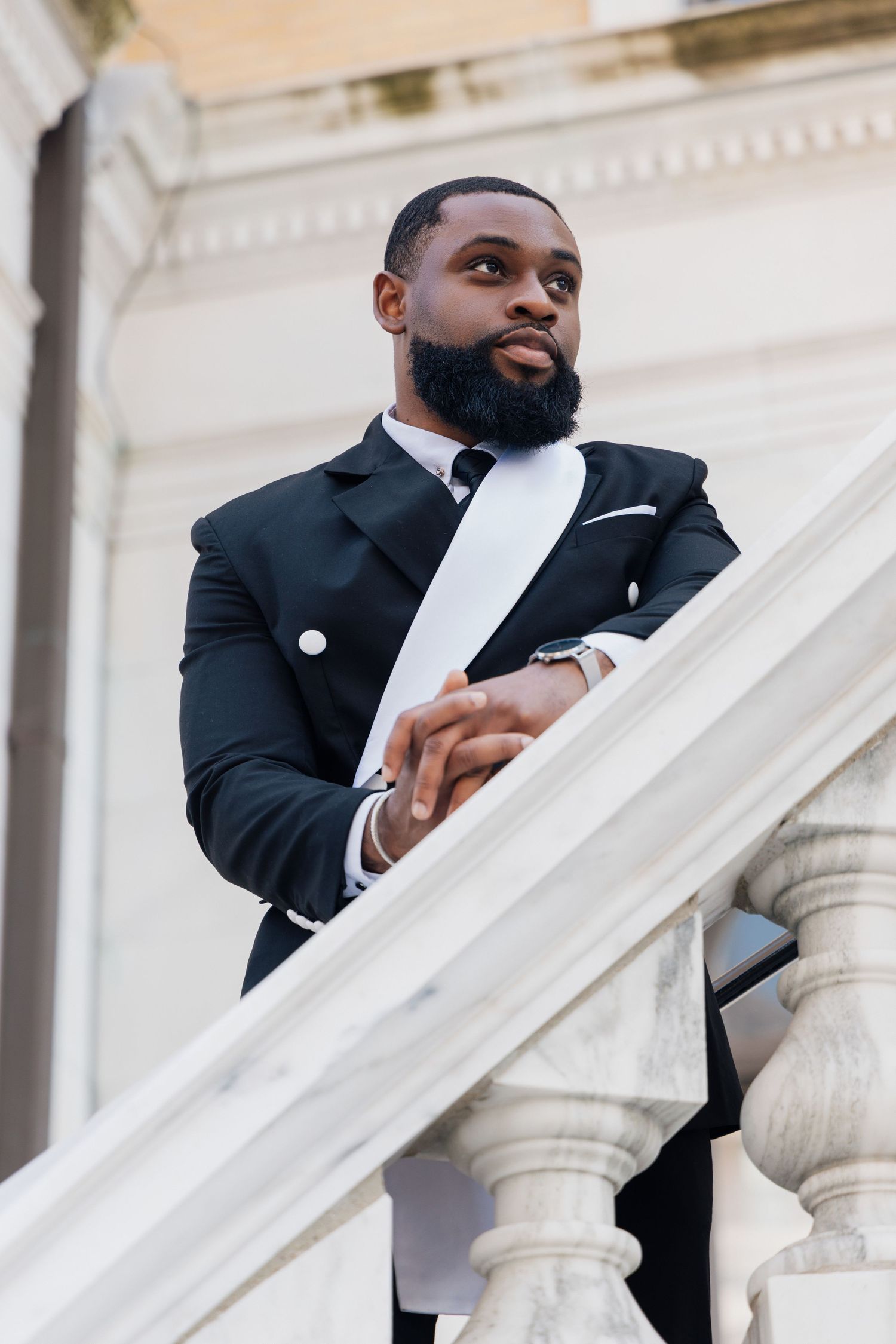 Elegant Black man in a tailored black tuxedo leans on marble staircase railing outdoors, exuding confidence.