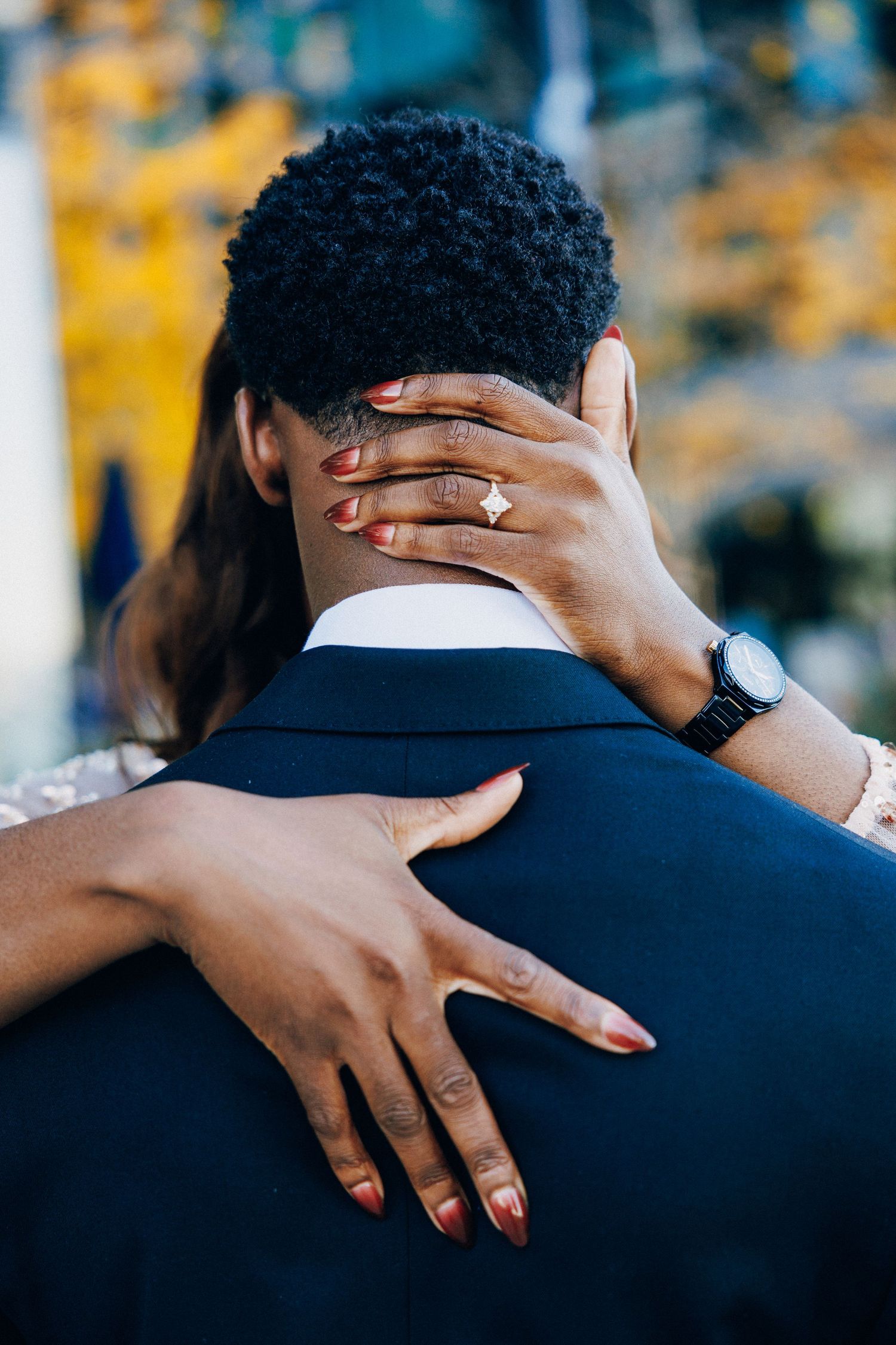 A woman with red nails and a gold engagement ring embraces a man in a navy suit from behind.