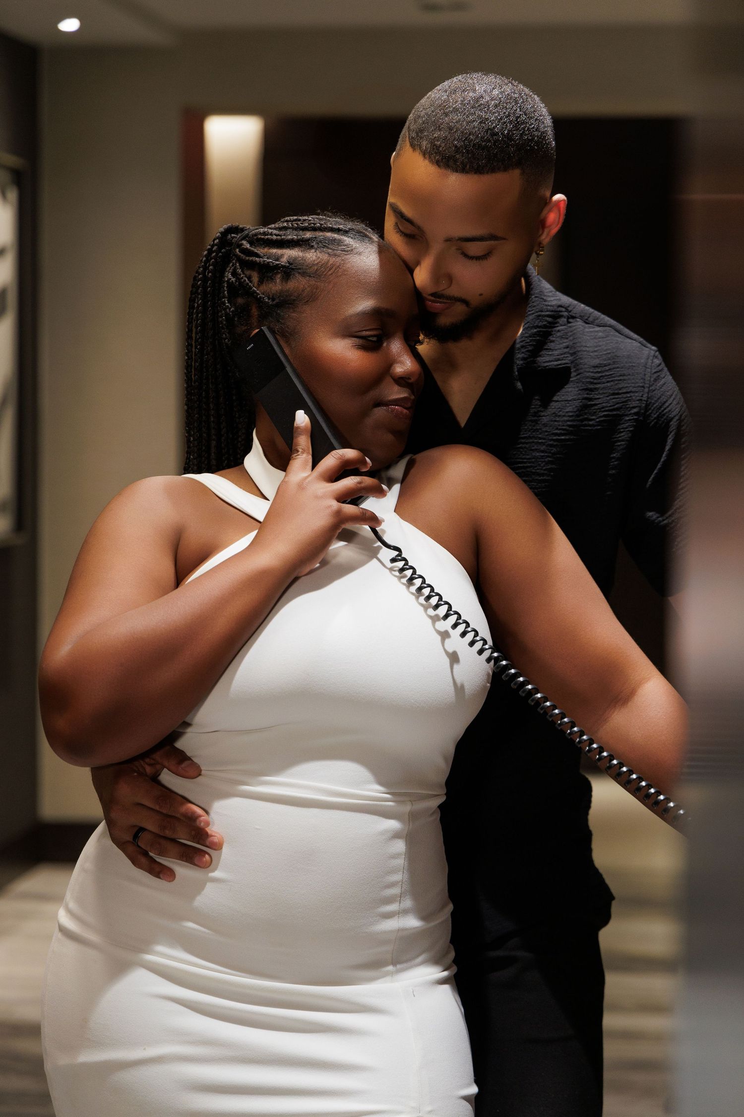 Couple embracing romantically, woman in white dress holding phone, man in black shirt, elegant indoor setting.