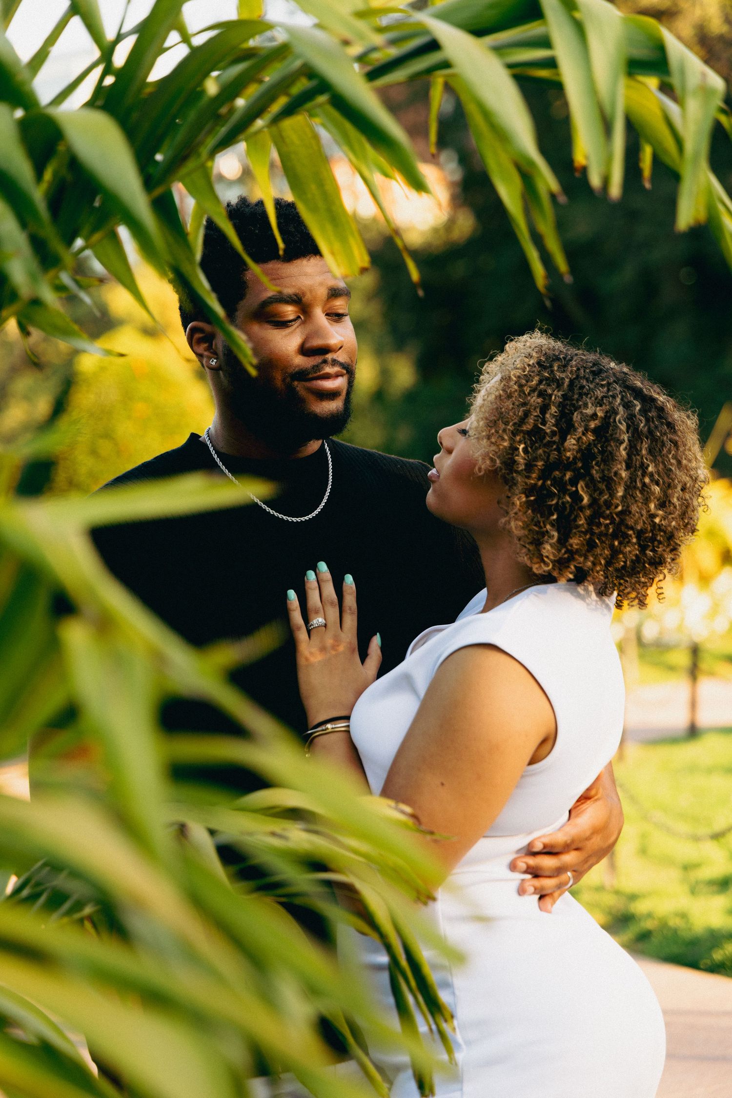 A couple embraces romantically among tropical palm leaves, the woman in white gazing up at her partner in a black shirt.