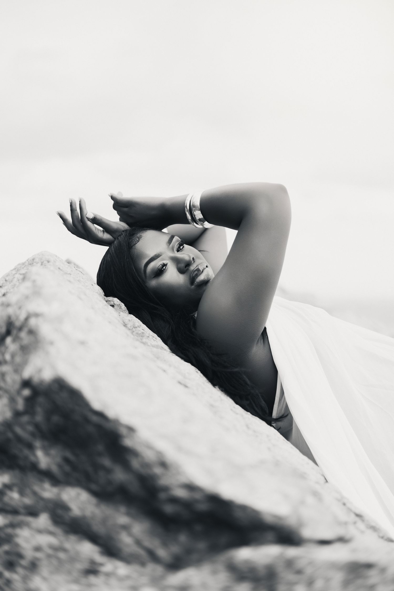 Black and white photo of a smiling woman reclining on rocks, wearing a white dress and bracelet, with arm raised gracefully.