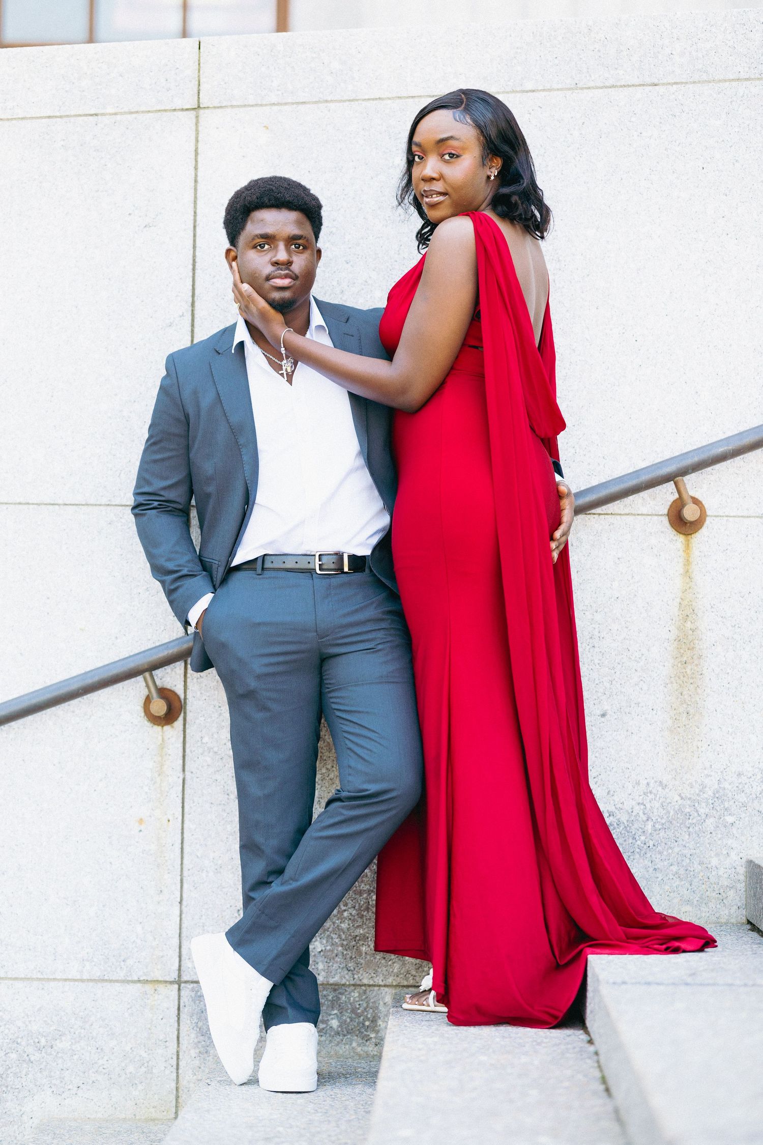 Stylish couple on outdoor steps, woman in flowing red gown touching mans face.