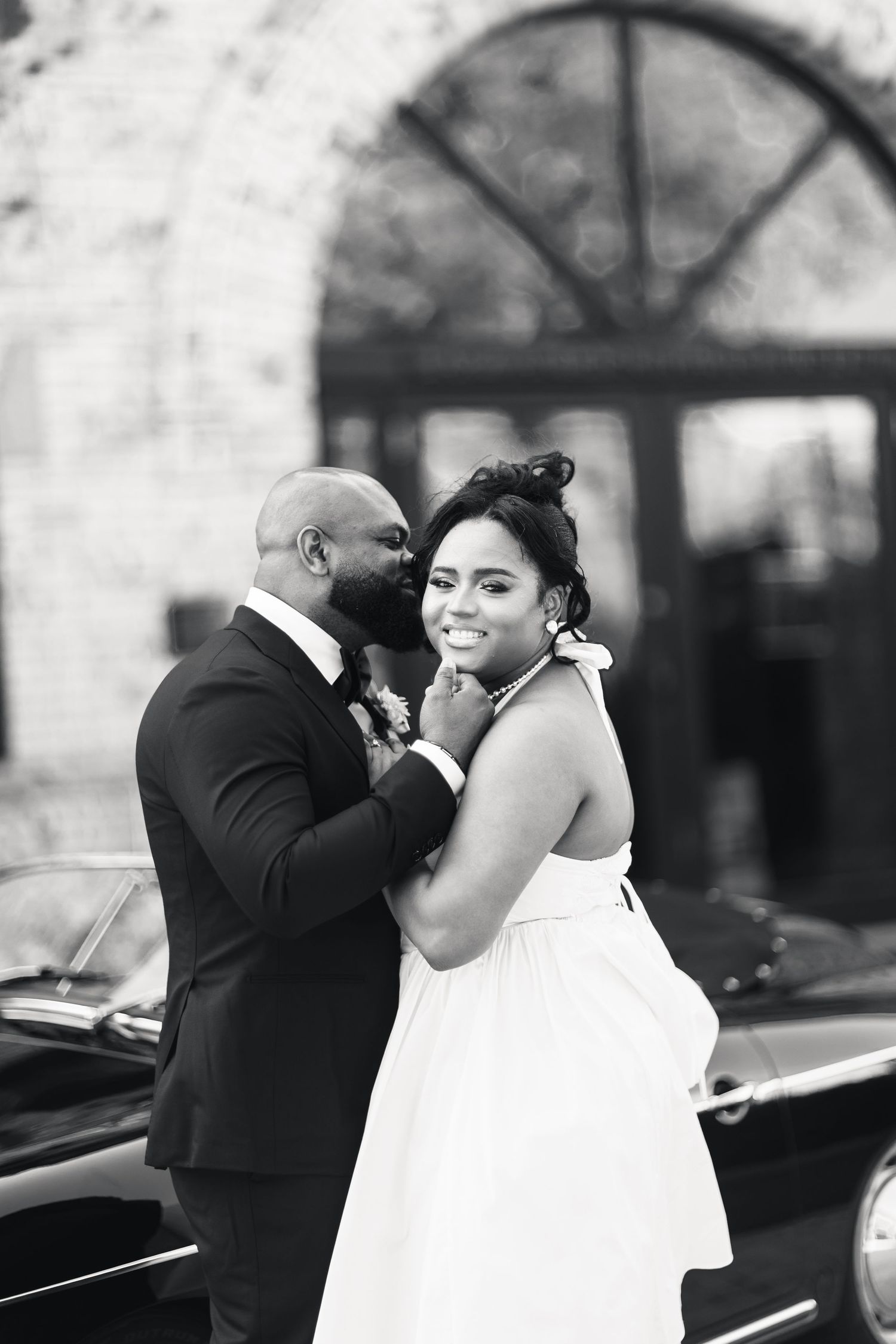 Black and white portrait of smiling bride and groom embracing beside a classic car at wedding venue.