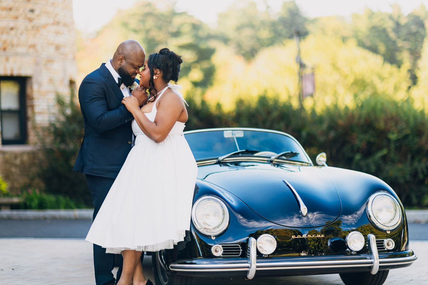 Newlywed couple sharing a kiss beside a classic vintage blue convertible car outdoors on their wedding day.
