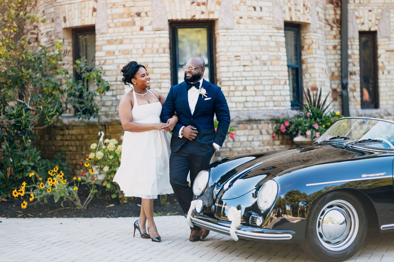 Bride and groom in wedding attire smile beside a vintage black Porsche outside a brick building.