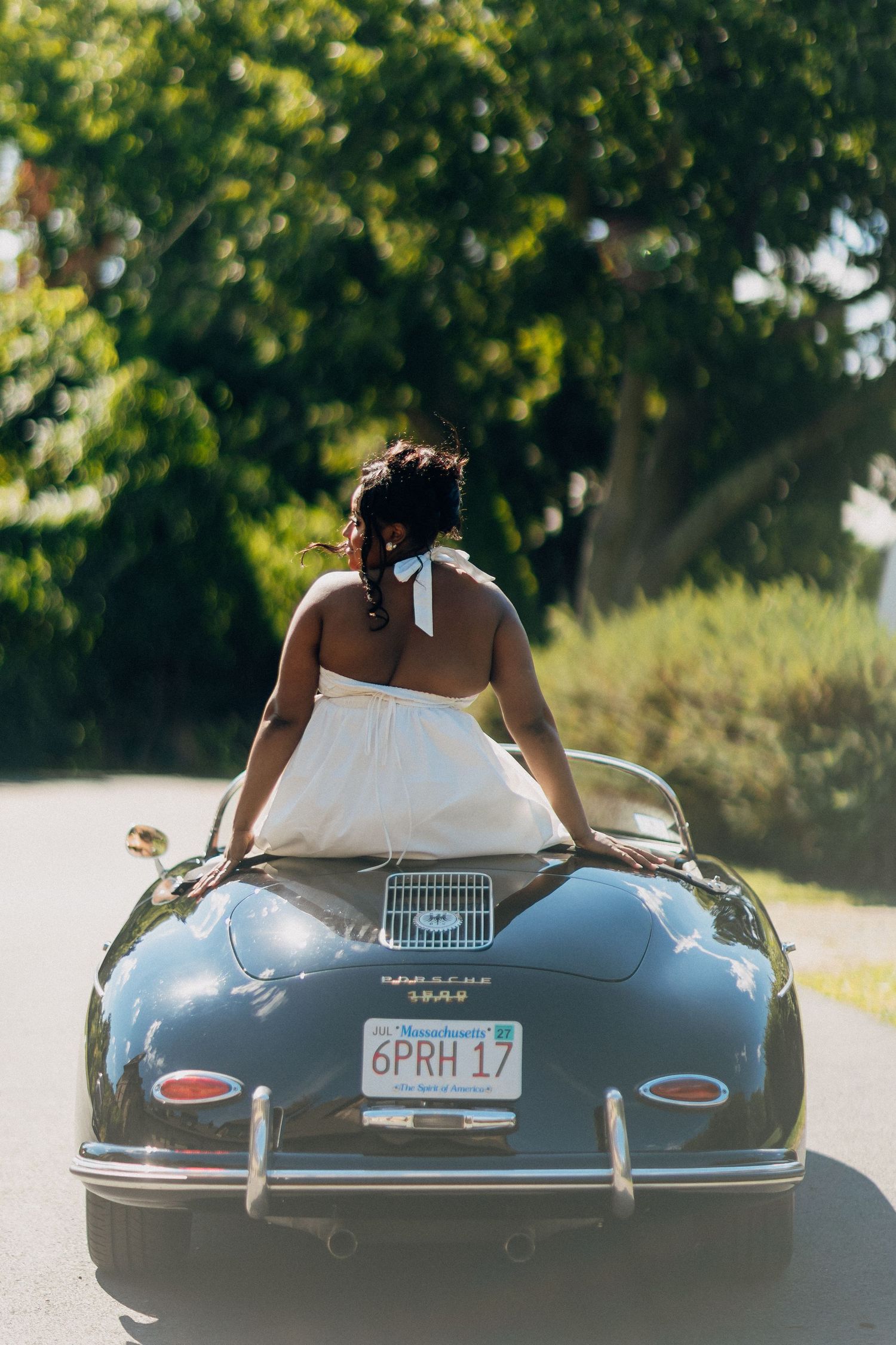 Bride in white halter dress leaning over the trunk of a vintage black Porsche 356 convertible.