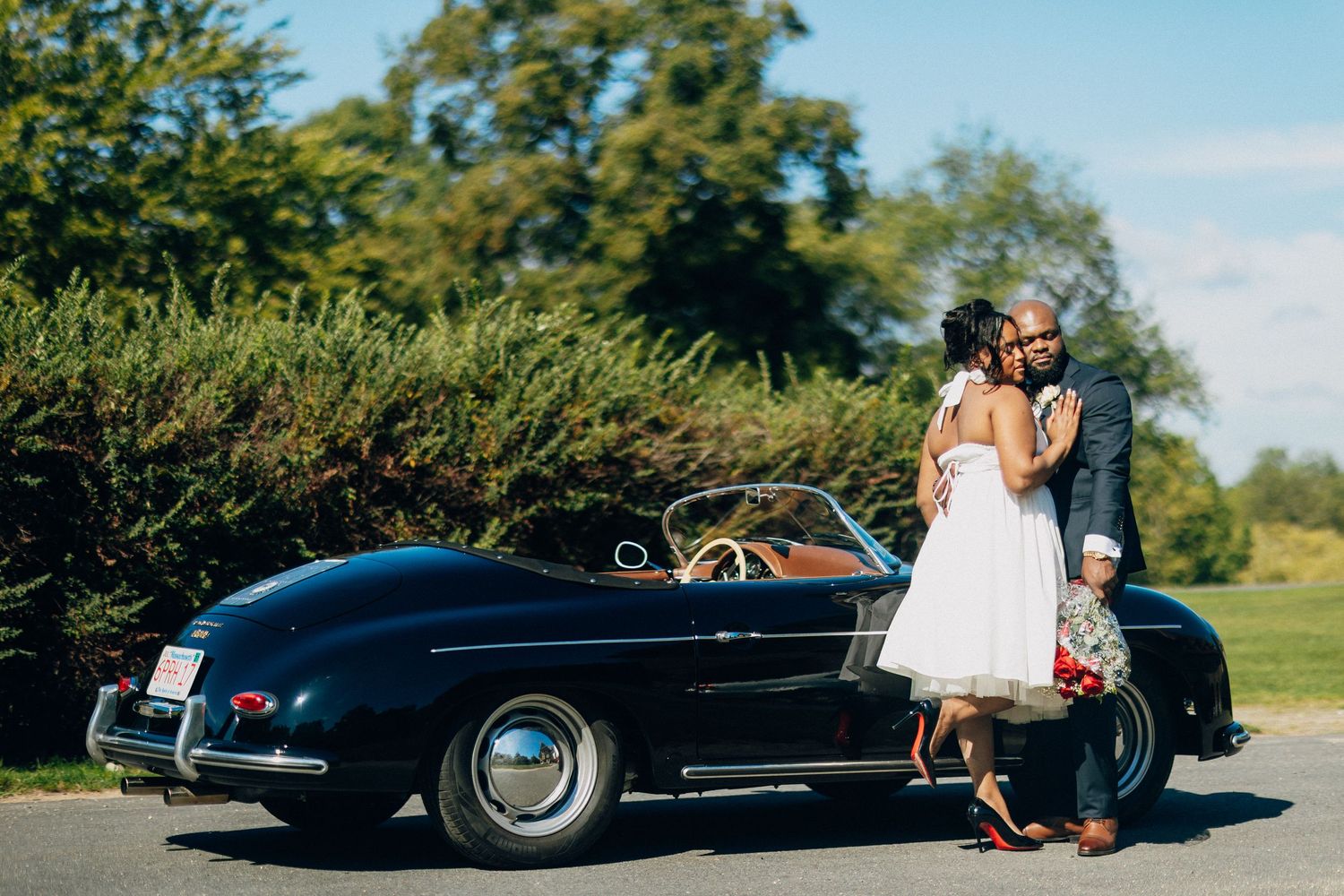 African American couple kissing beside a classic black Porsche convertible on a sunny wedding day.