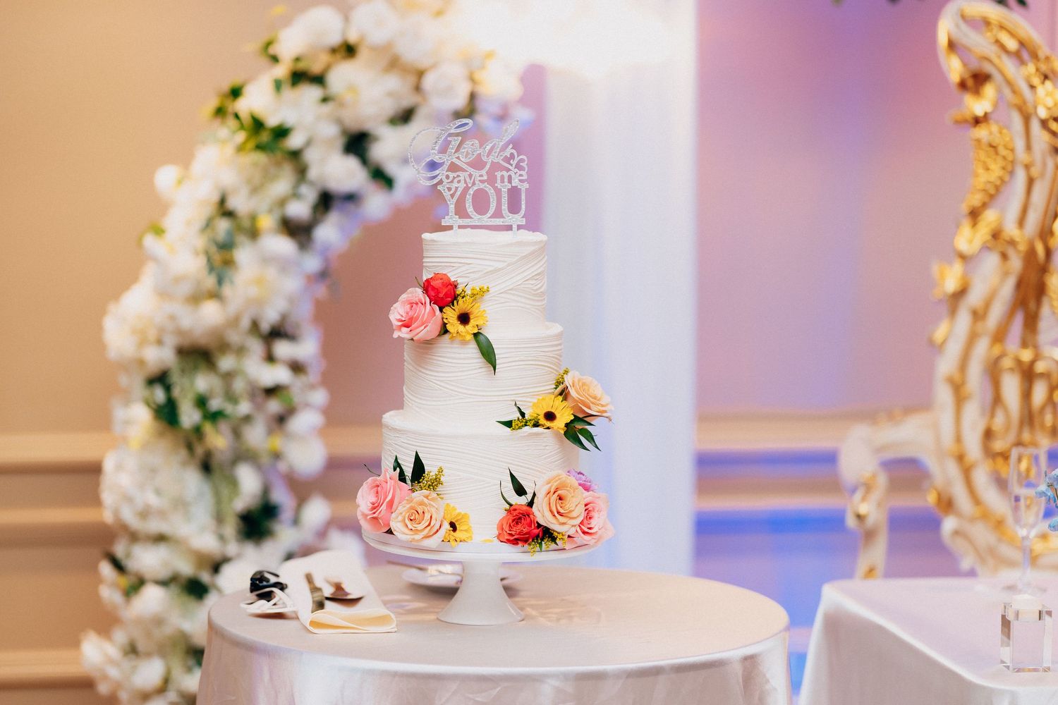 Elegant three-tier white wedding cake adorned with colorful flowers and a decorative topper, displayed at a reception.