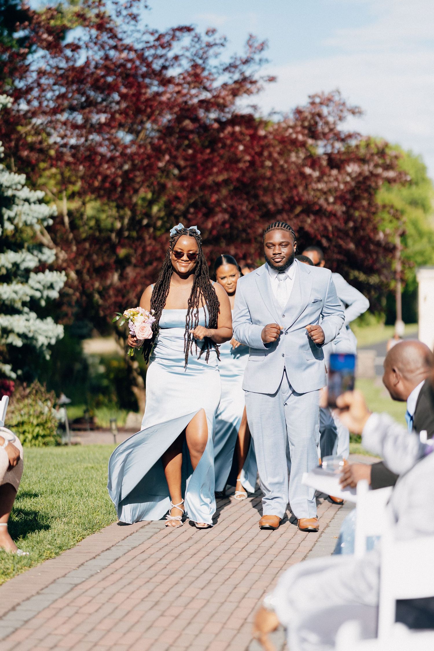 Bridesmaid in light blue dress and groomsman in blue suit walking down brick wedding aisle.