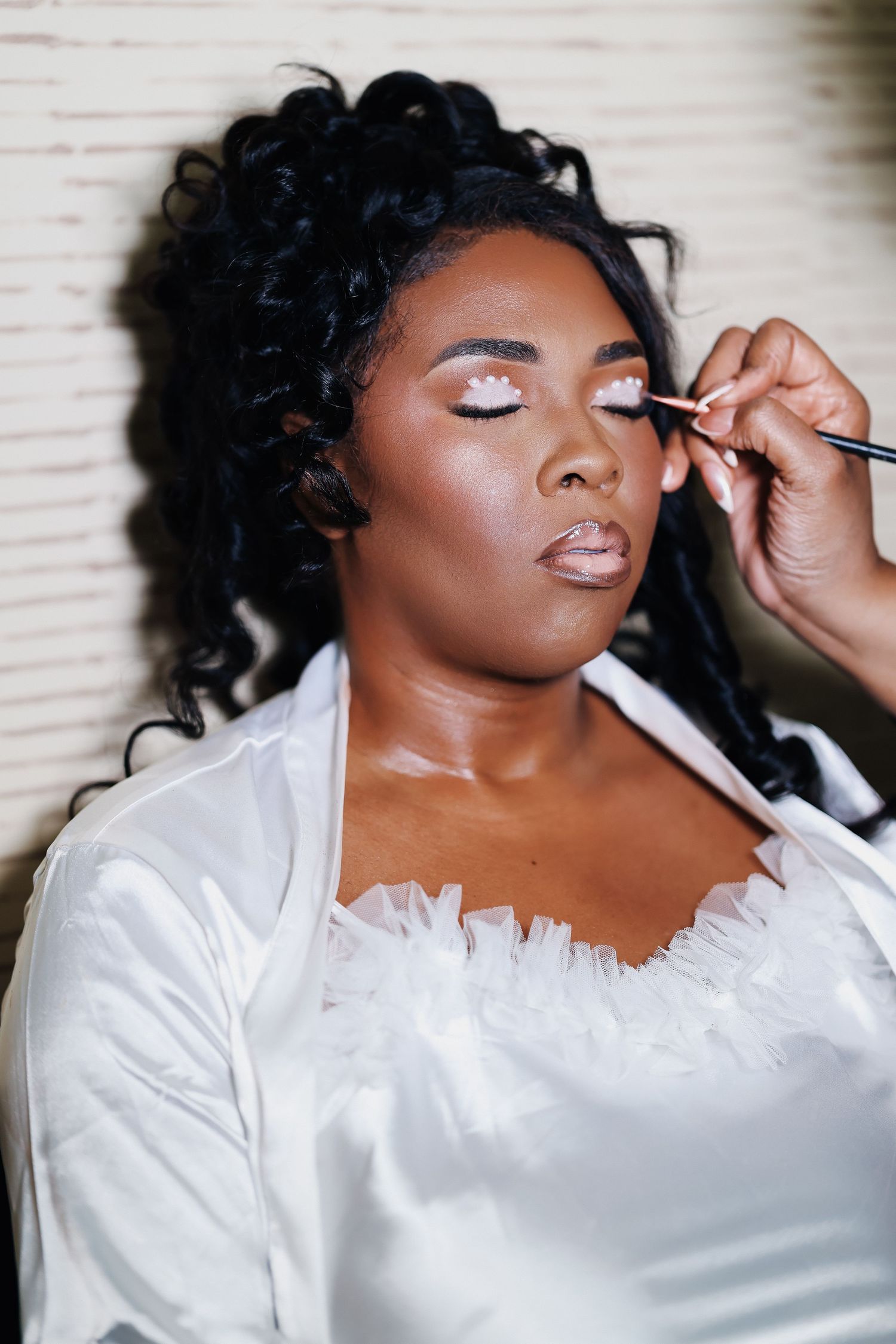 African American bride getting eye makeup applied by artist while wearing white satin robe before wedding ceremony.