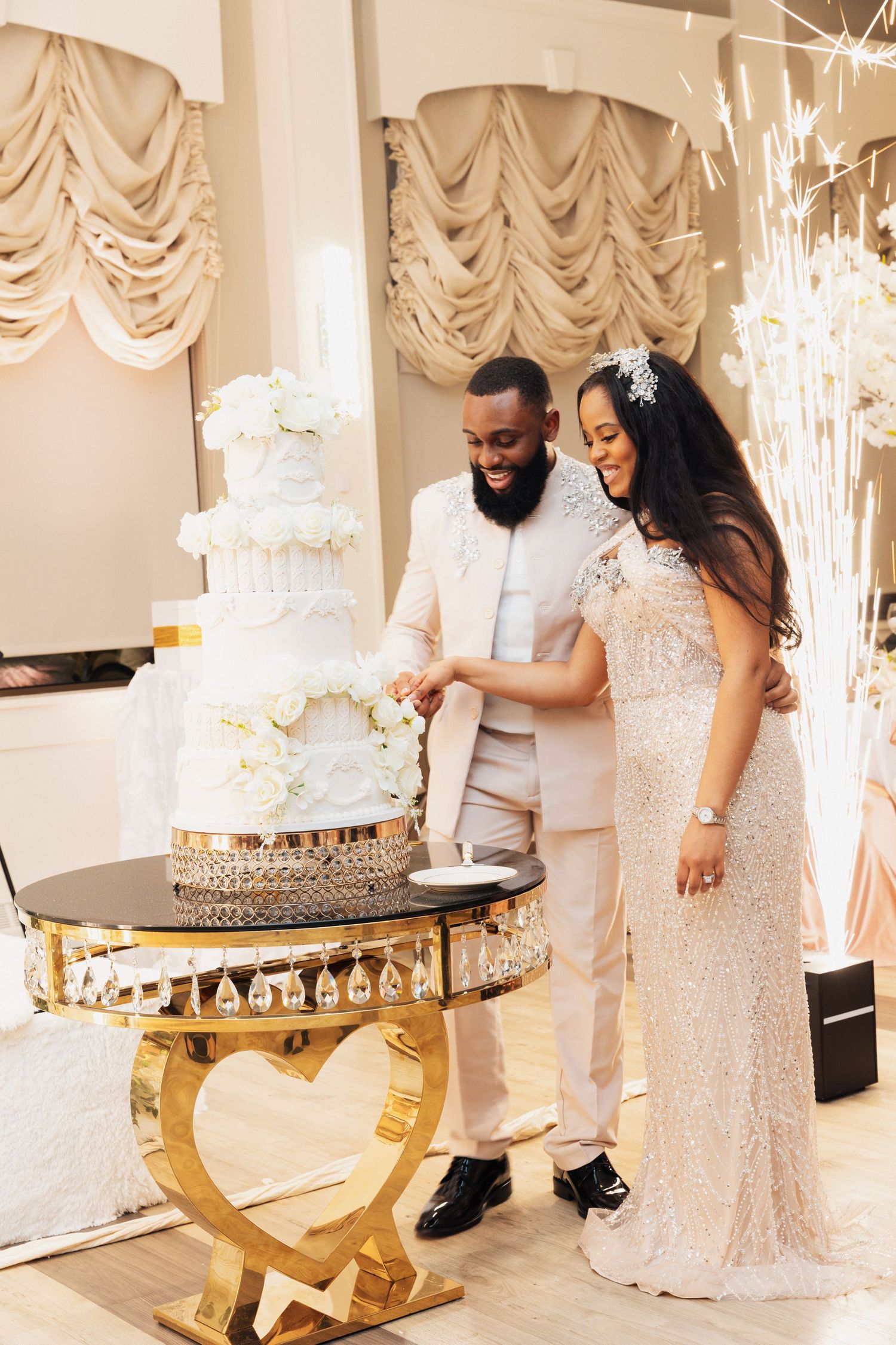 Couple cutting a white tiered wedding cake on a gold heart-shaped table in an elegant reception venue.