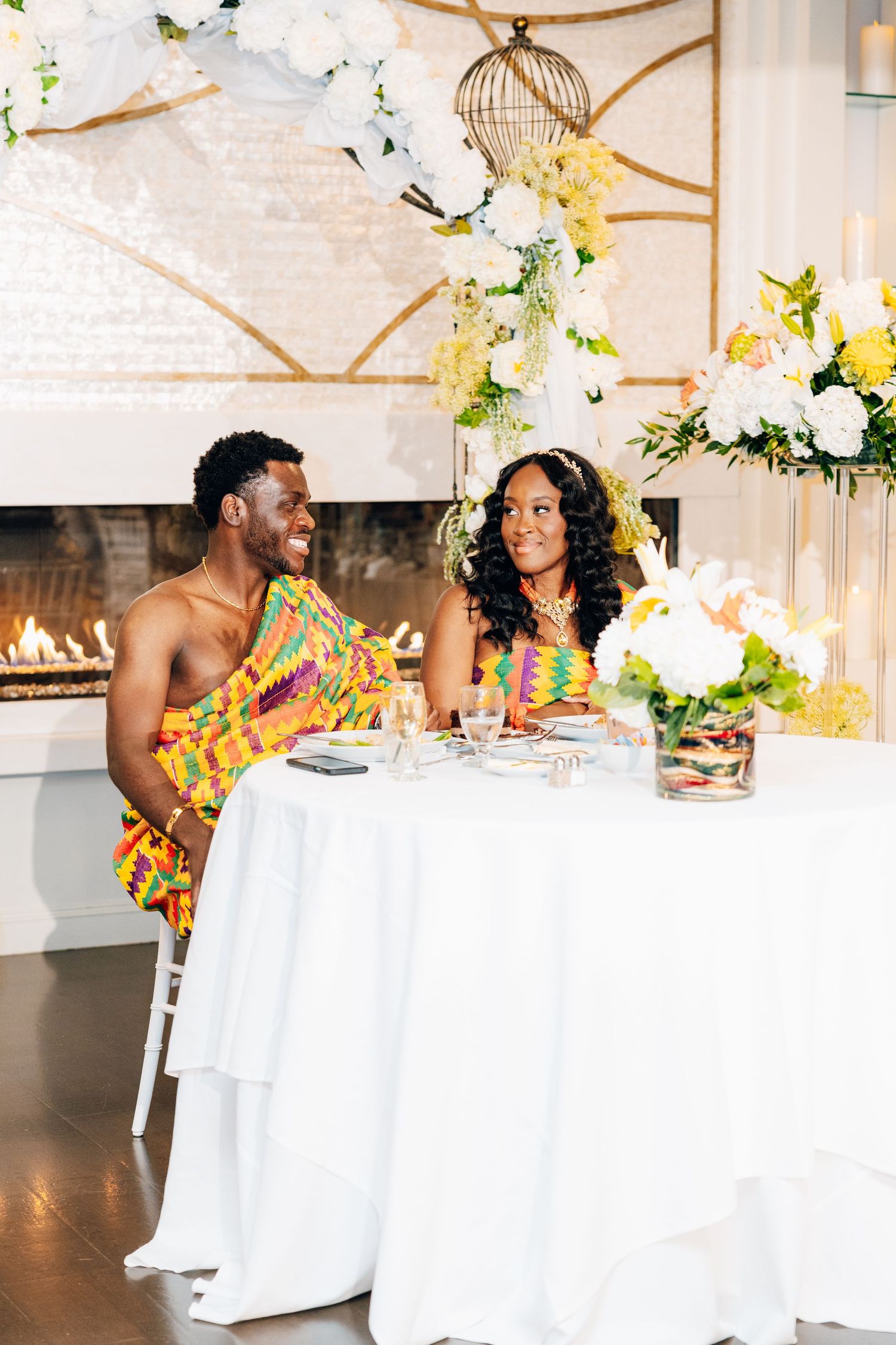 Couple in colorful Kente cloth attire seated at elegant white-draped sweetheart table with floral arrangements.