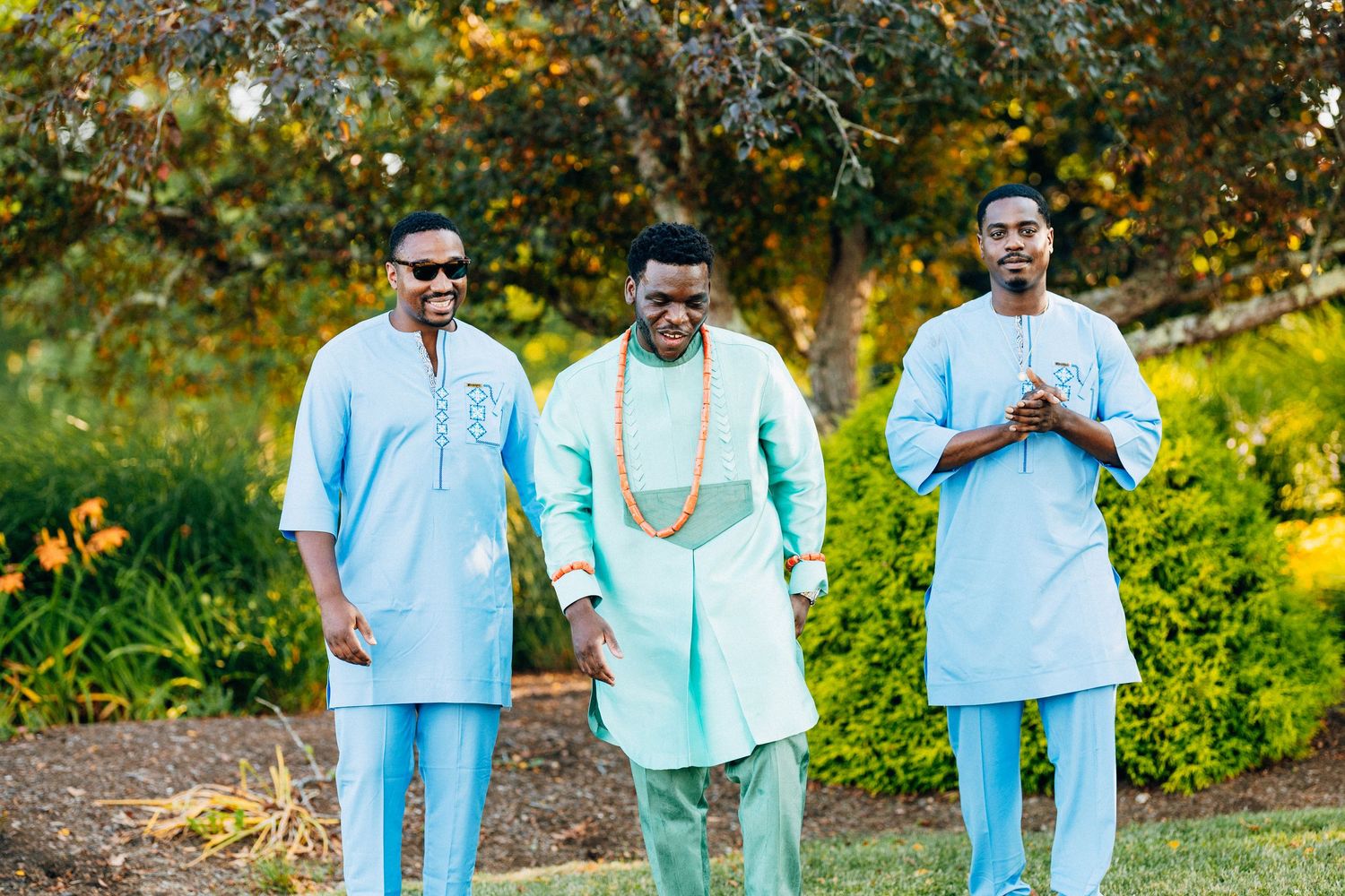 Three men in matching light blue traditional African attire walk outdoors at a wedding celebration.
