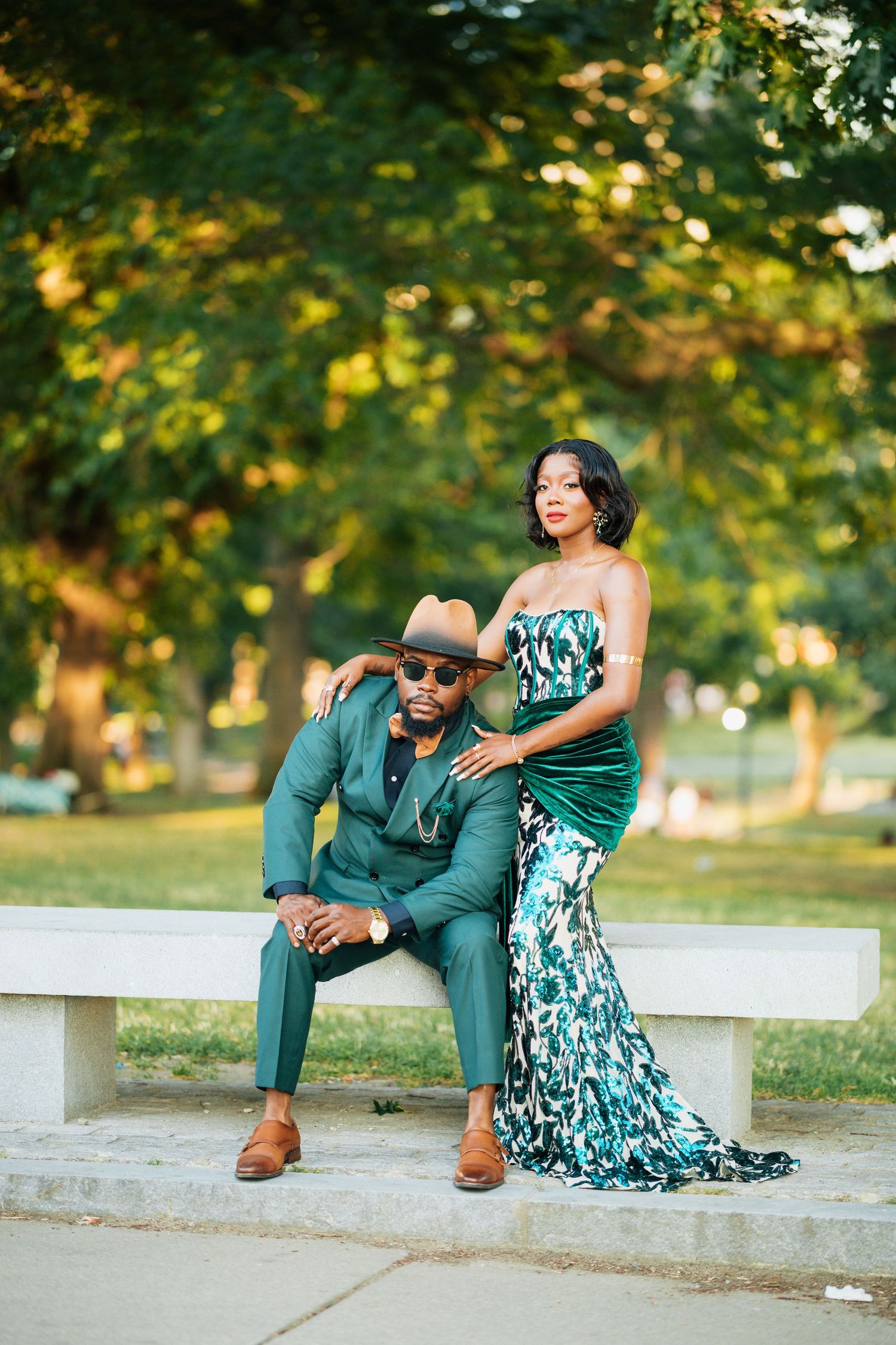 Elegant couple in matching green outfits posing in a lush park, man seated in suit and hat, woman in floral gown.