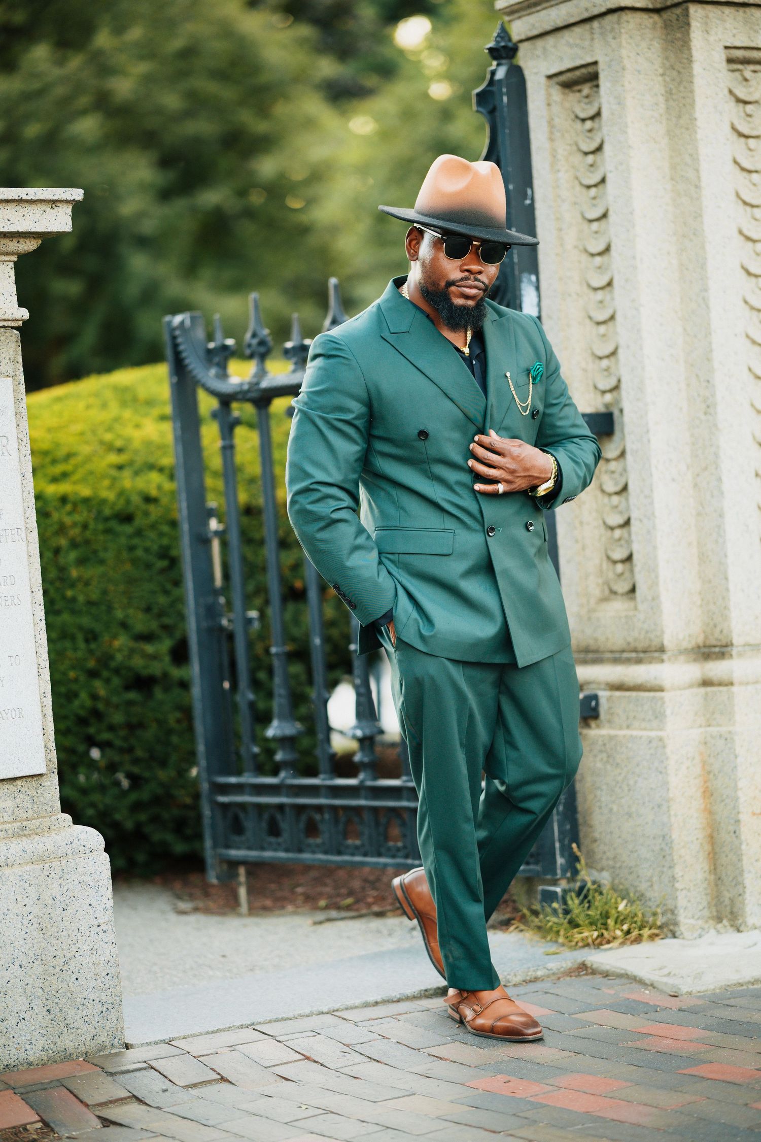 Dapper man in emerald green suit and tan hat posing by ornate iron gate.