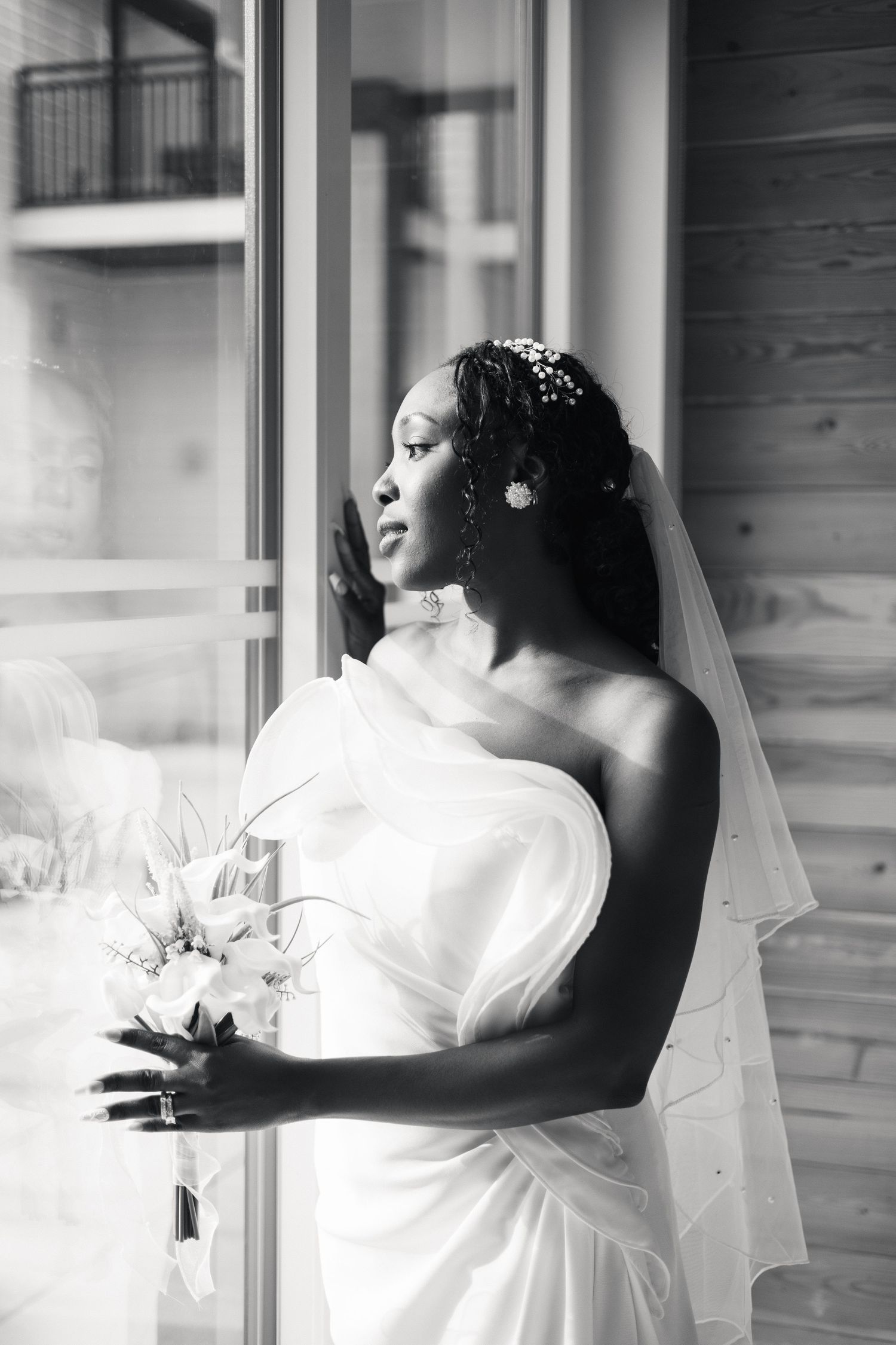Elegant Black bride in white off-shoulder gown and veil gazing out window, holding white floral bouquet.