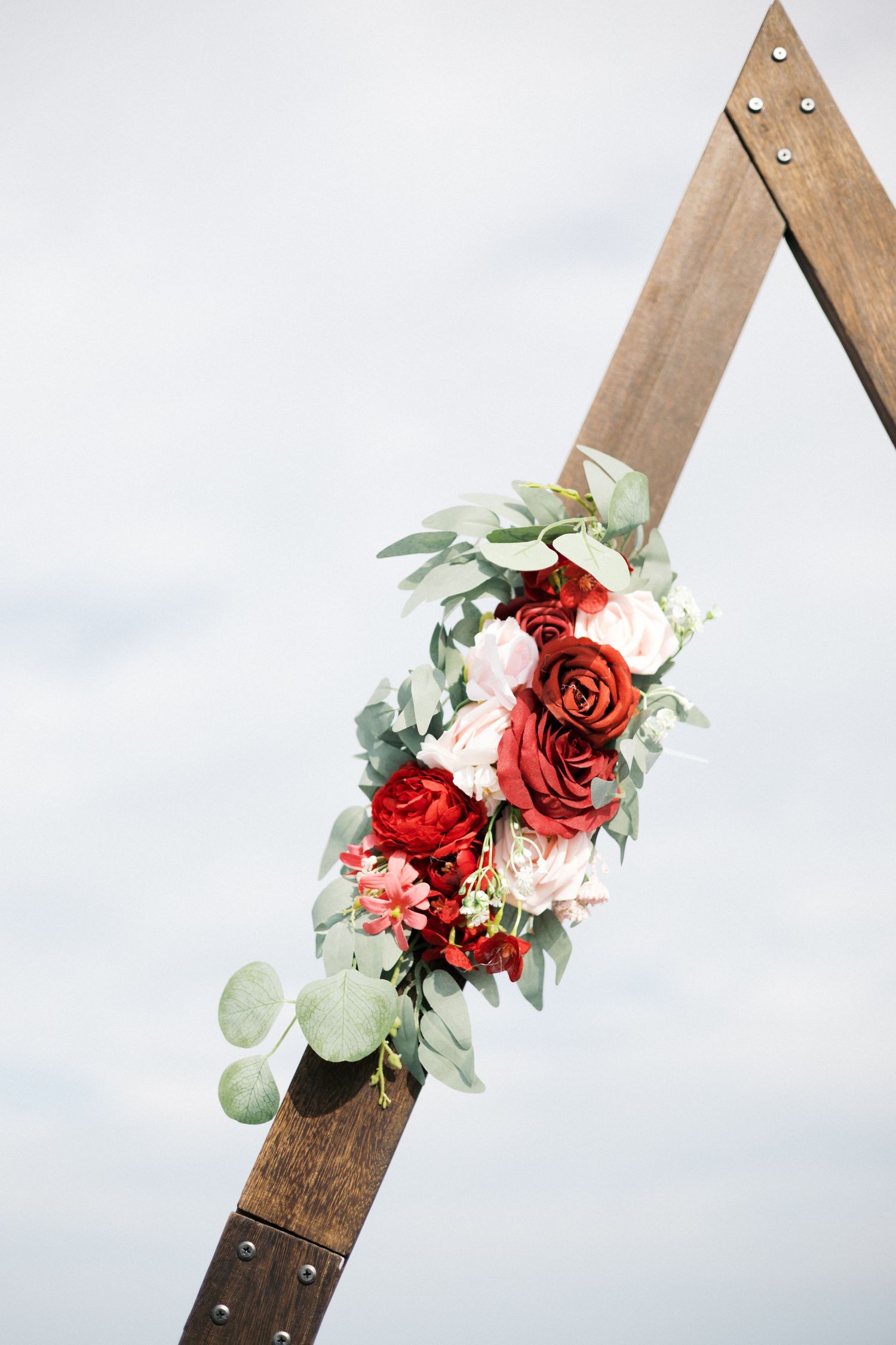 Rustic wooden wedding arch adorned with red and blush roses and eucalyptus greenery against a soft gray sky.