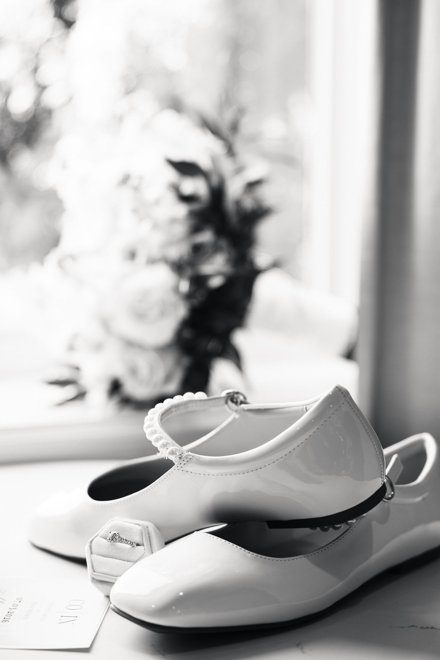 Black and white photo of bridal flat shoes with pearl strap, engagement ring, and blurred floral bouquet by window.