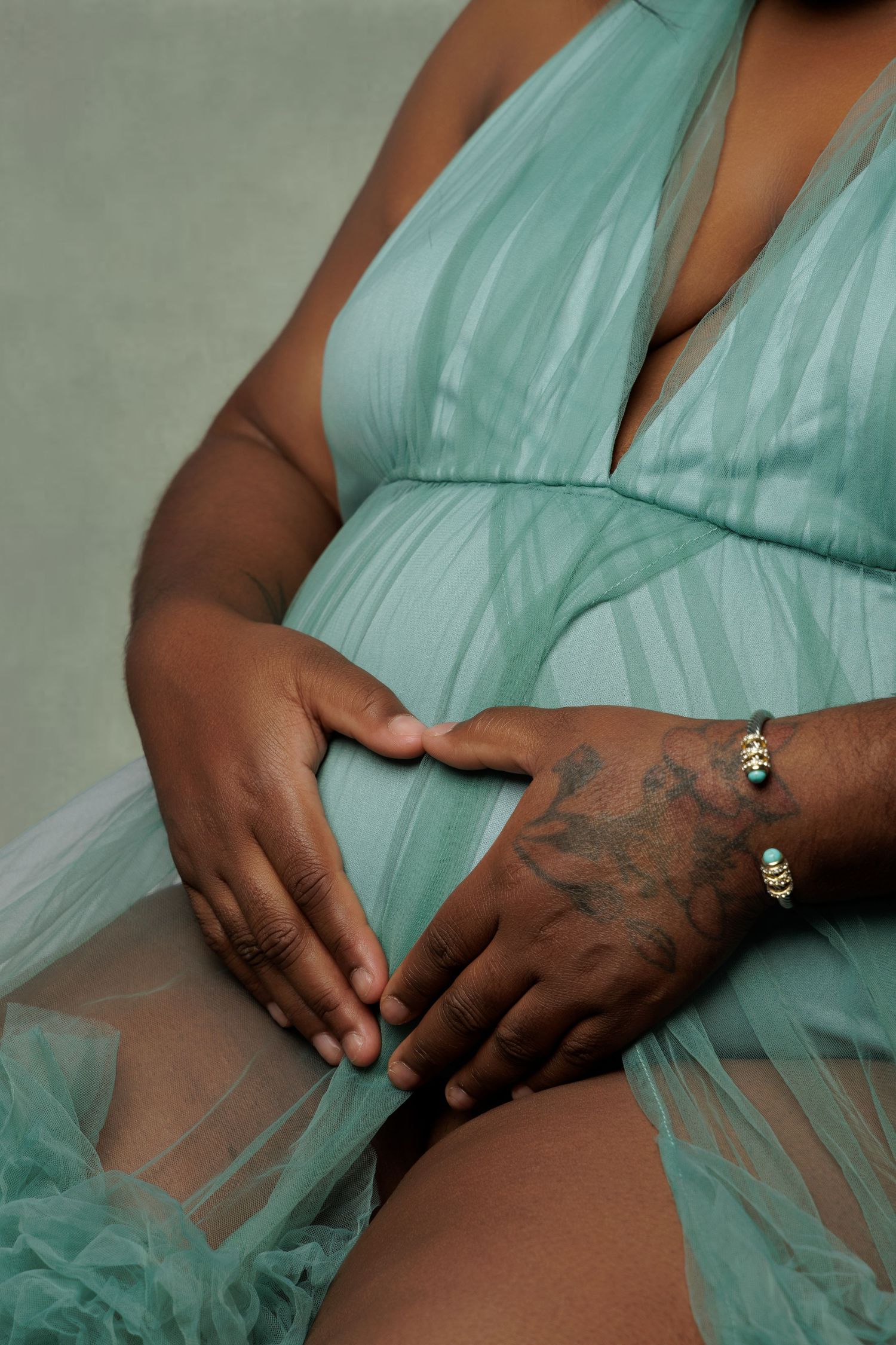Pregnant woman in mint green tulle dress cradling baby bump, with tattooed hands and gold bracelet visible.