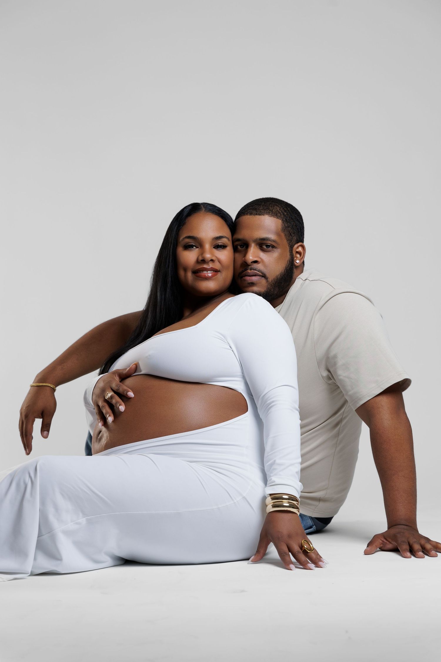 A pregnant woman in white sits with her partner behind her, both smiling in a tender maternity photo shoot.